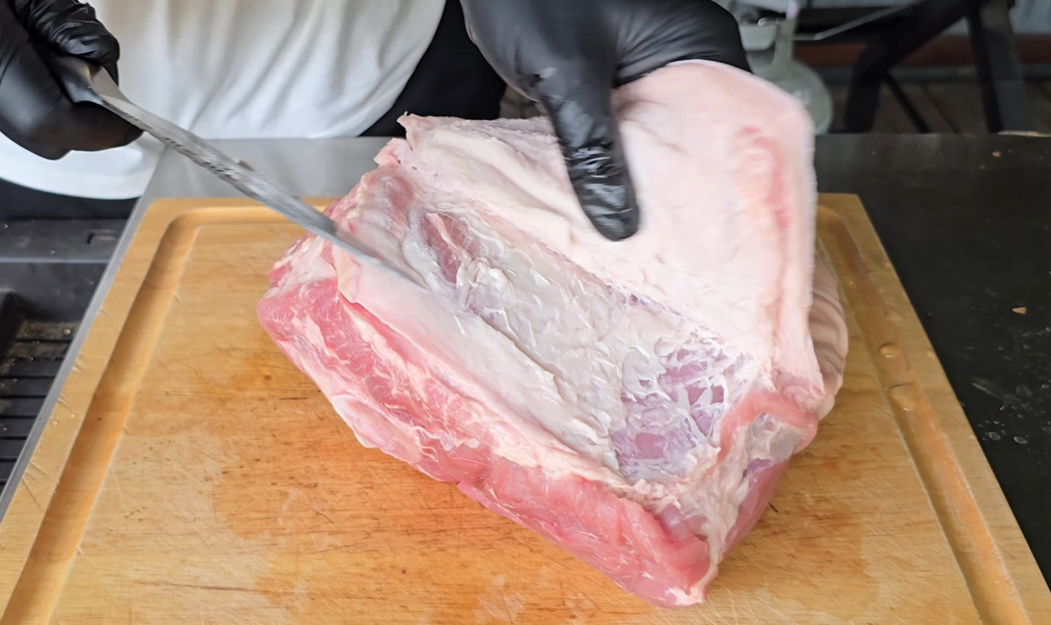 A person wearing black gloves is trimming fat from a large raw pork roast on a wooden cutting board using a knife.