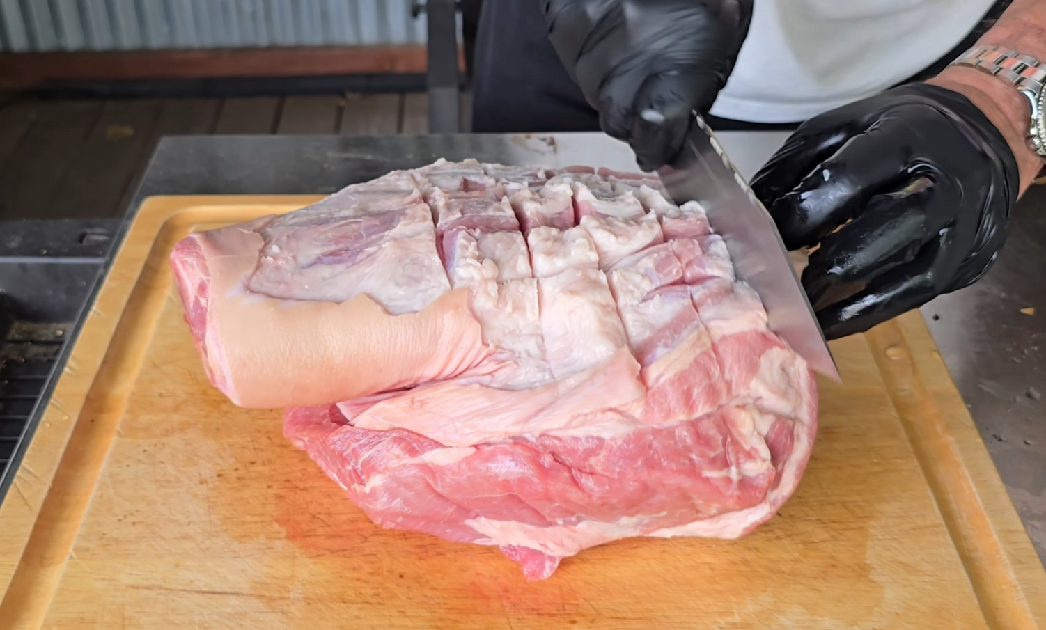 A person wearing black gloves scores the fat on a large, raw pork shoulder with a knife on a wooden cutting board.