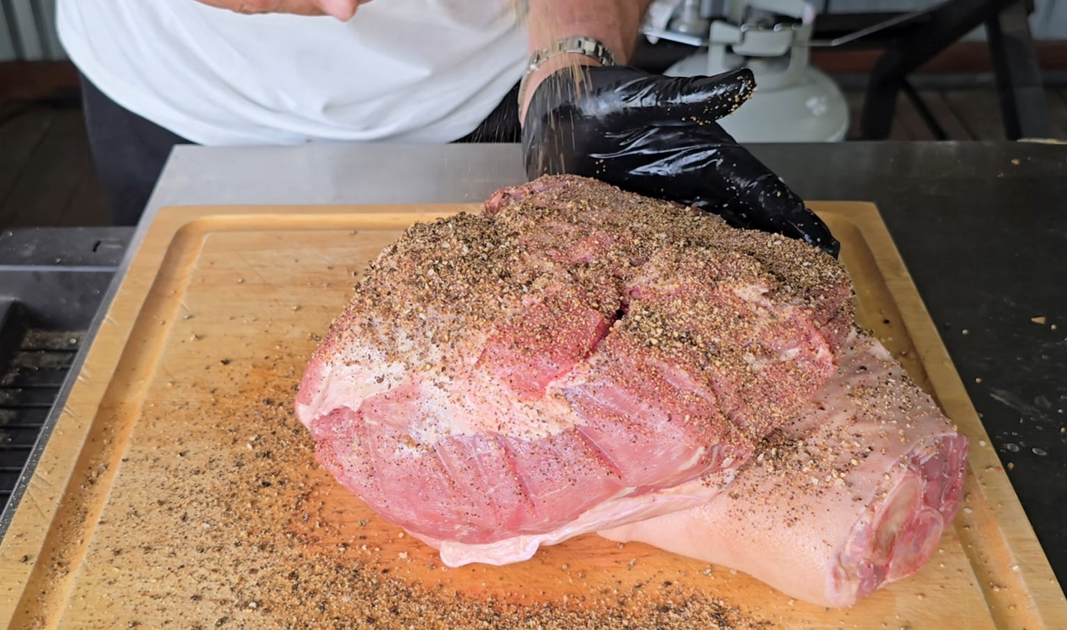 A person wearing a black glove seasons a large raw cut of meat on a wooden cutting board with pepper and spices.