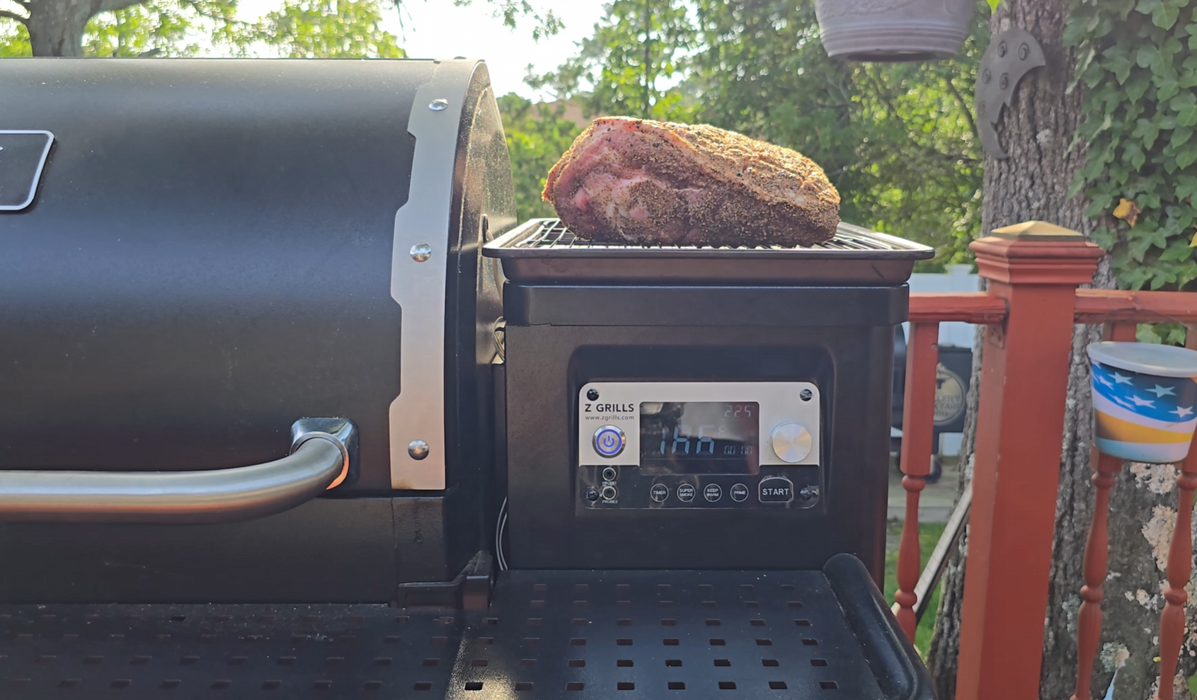 A large, seasoned cut of meat sits on a rack above a smoker grill with a digital control panel, outdoors on a sunny day, with trees and a flag-themed cup in the background.