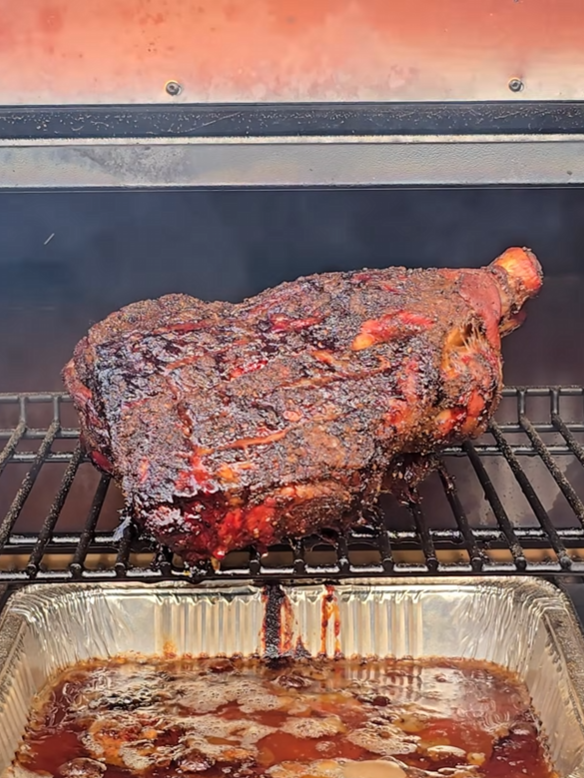 A large, seasoned piece of meat is smoking on a grill, with juices dripping into a foil pan below. The meat has a dark, crusty coating and the grill is inside a smoker.