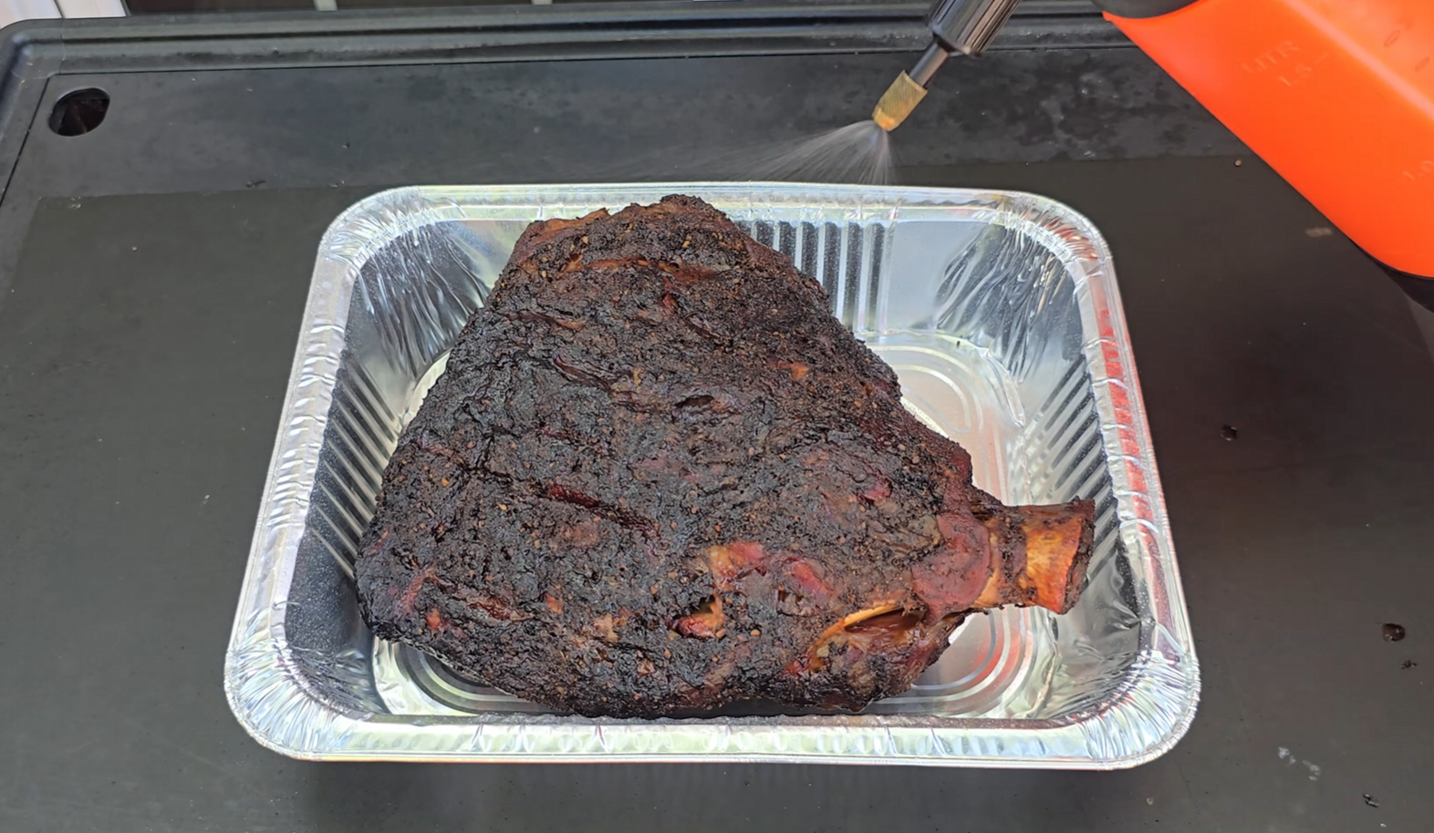 A large piece of smoked meat with a dark, crispy crust sits in a disposable aluminum tray. A hand holds a spray bottle, misting the meat. The scene appears to be part of a barbecue or outdoor cooking setup.