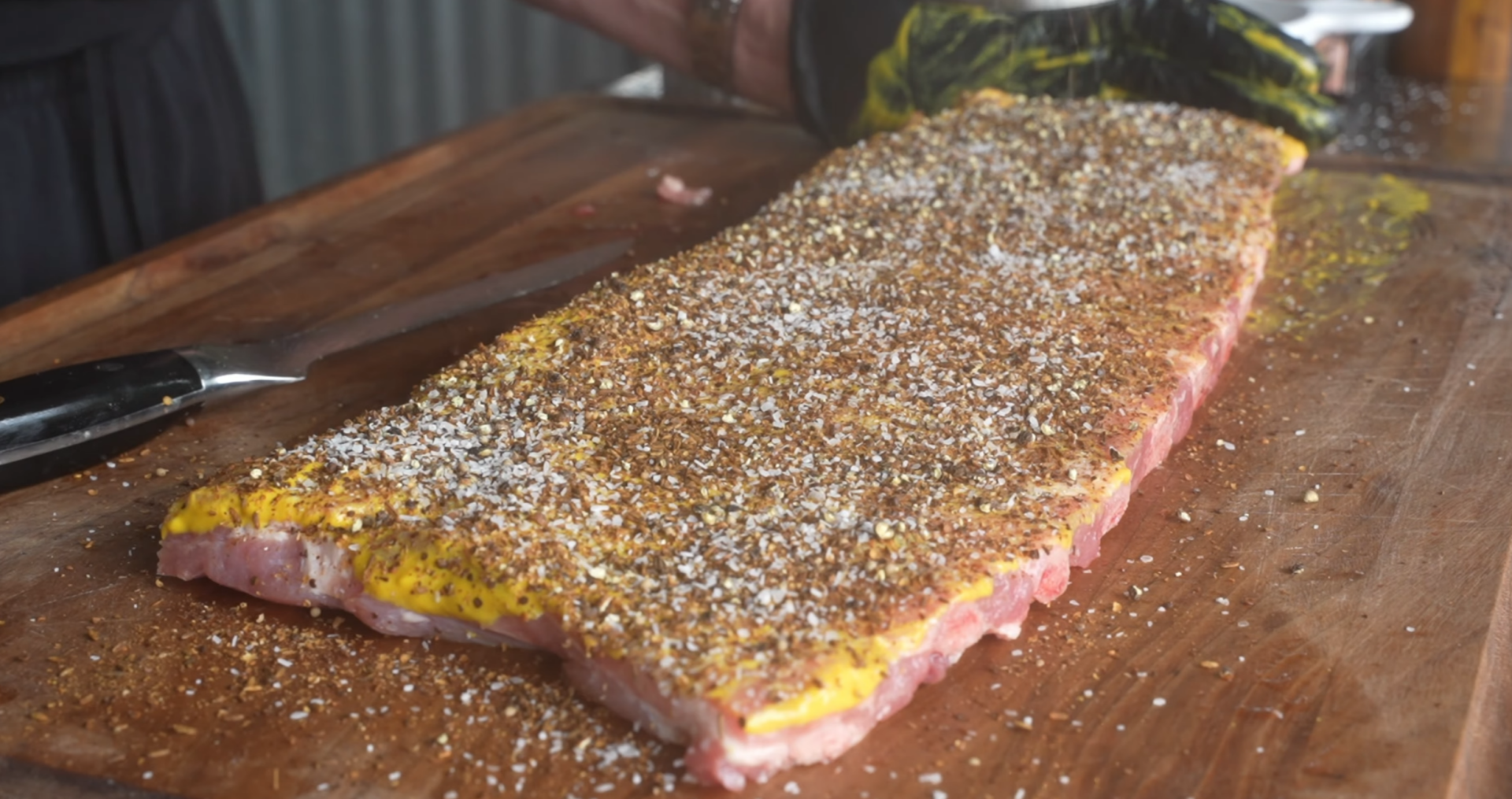 A slab of raw pork ribs on a wooden board, coated with yellow mustard and sprinkled heavily with seasoning. A gloved hand is visible applying the seasoning. A knife lies next to the ribs.