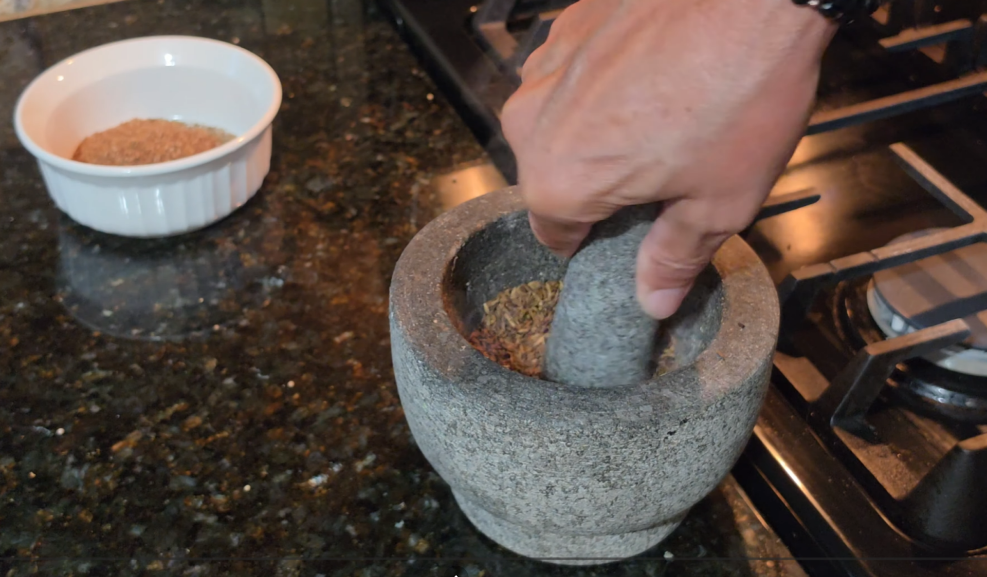A hand uses a stone mortar and pestle to grind spices on a black granite countertop, with a small white bowl of spices nearby.