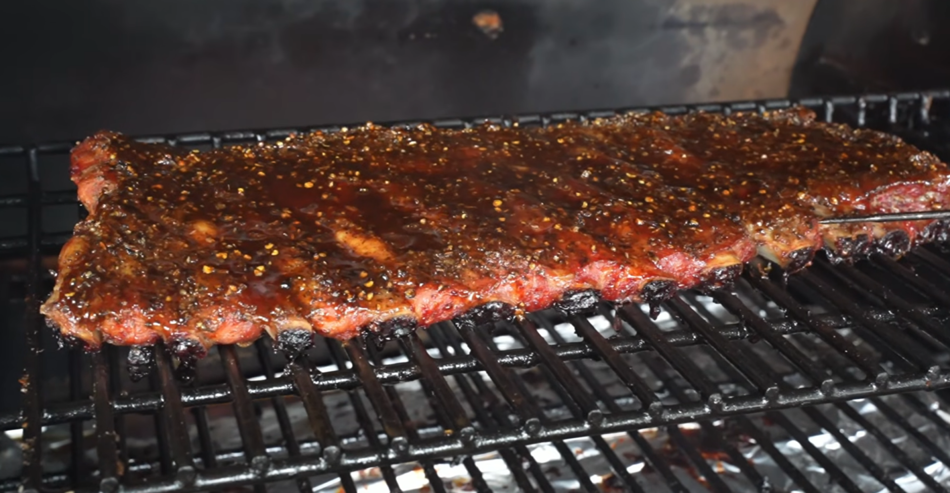 A rack of glazed ribs cooks on a grill, with a meat thermometer inserted on the right side, above glowing coals.