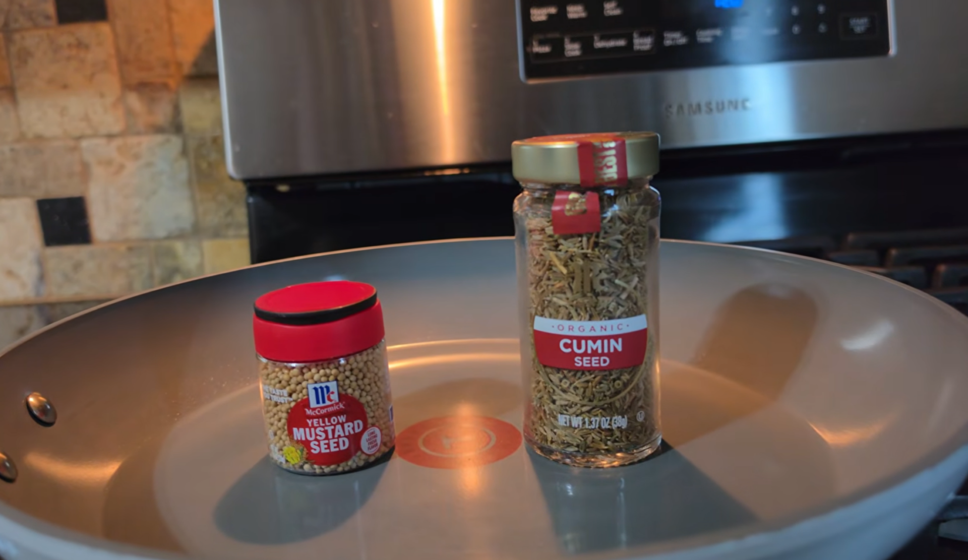 A jar of yellow mustard seed and a jar of organic cumin seed are placed on a gray frying pan in a kitchen with a stove and tiled backsplash in the background.