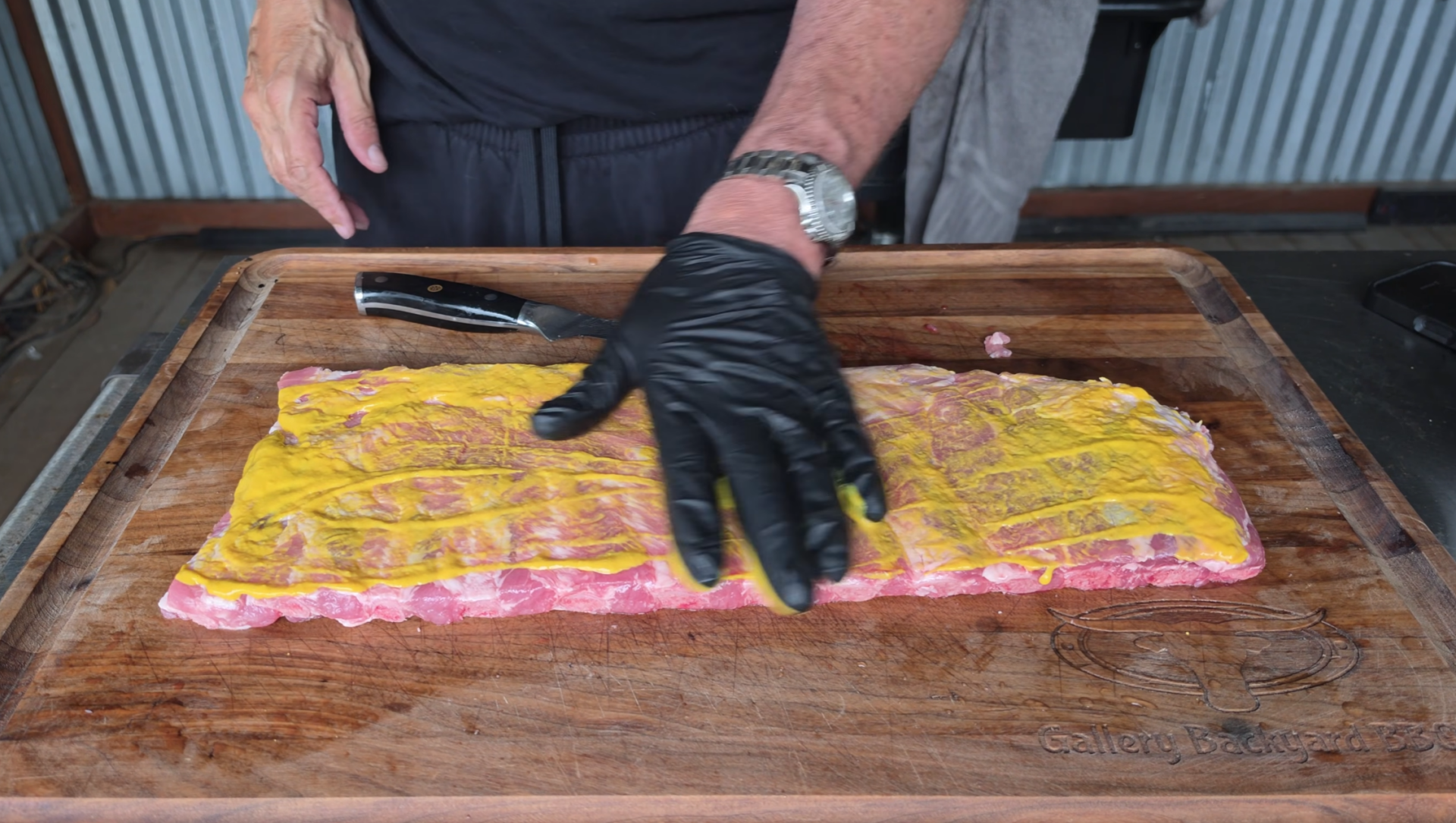 A person wearing black gloves spreads yellow mustard on a rack of raw ribs placed on a wooden cutting board. A knife is next to the meat.