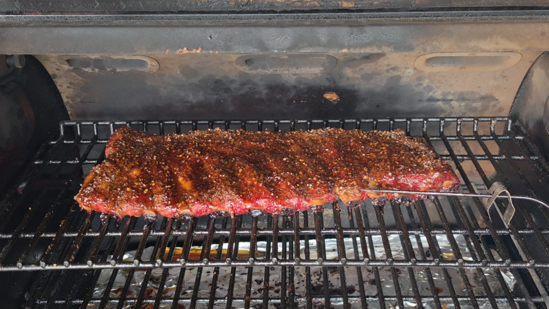 A rack of seasoned ribs is cooking on a grill, with a meat thermometer probe inserted on the right side. The grill grate is above a drip tray lined with foil. The ribs are browning and appear to be slow-cooked.