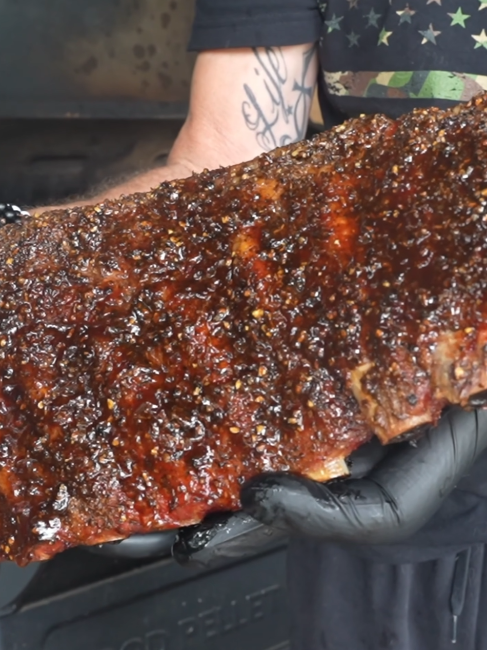 A person wearing black gloves holds a large, glazed, and seasoned rack of barbecued ribs in front of a grill.
