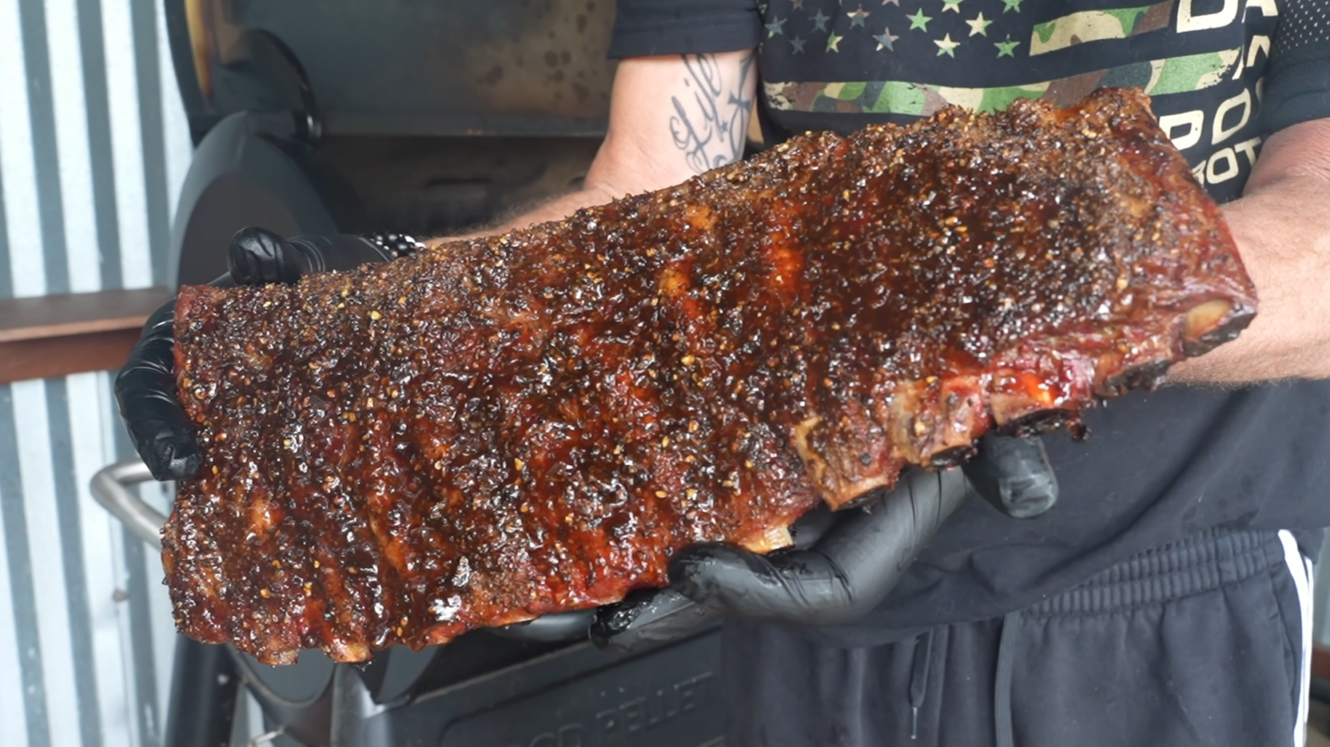A person wearing black gloves holds a large, glistening rack of barbecued ribs in front of a grill.