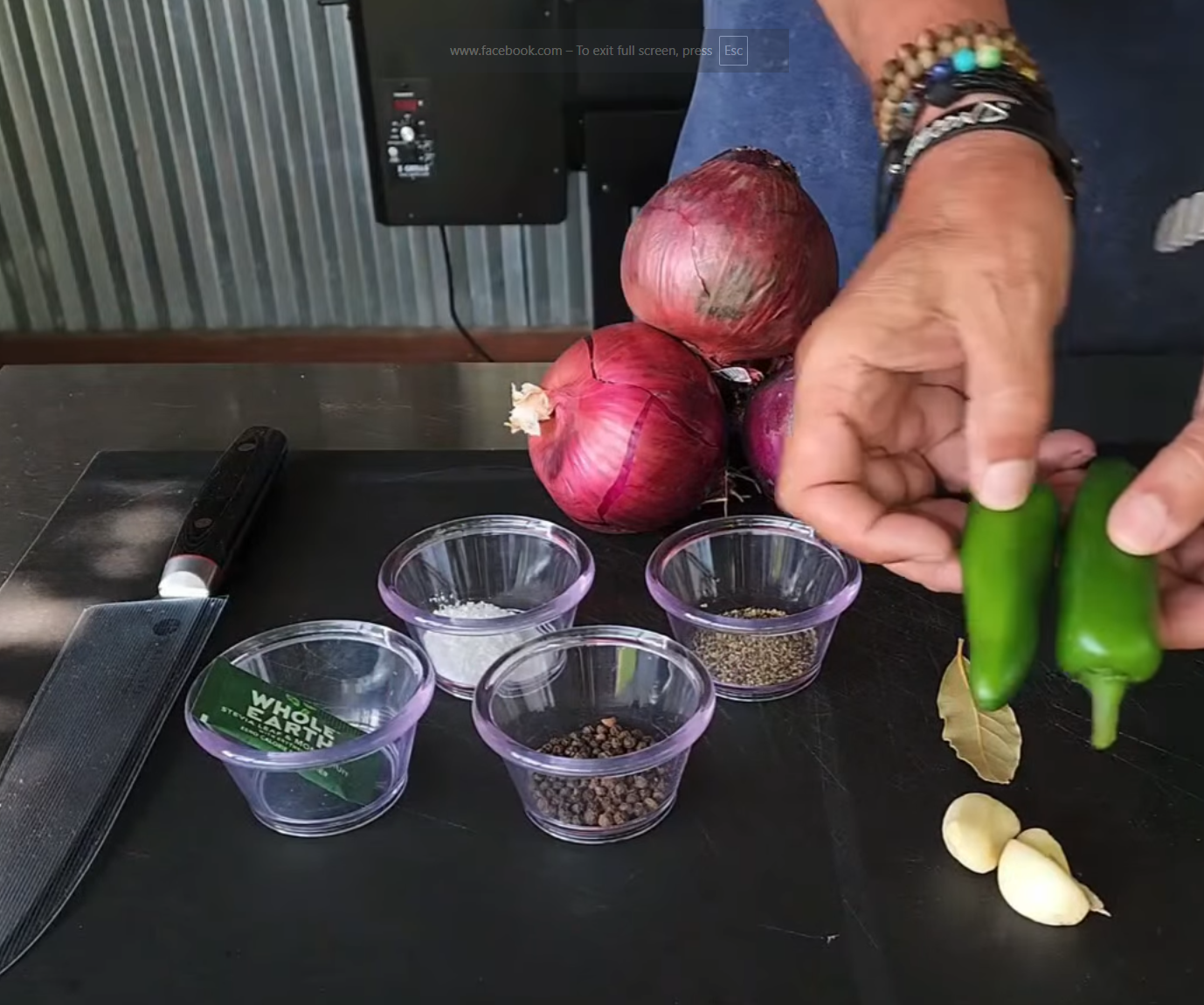 A person holds two green chili peppers next to red onions, garlic cloves, bay leaves, spices in small bowls, and a knife on a black cutting board.