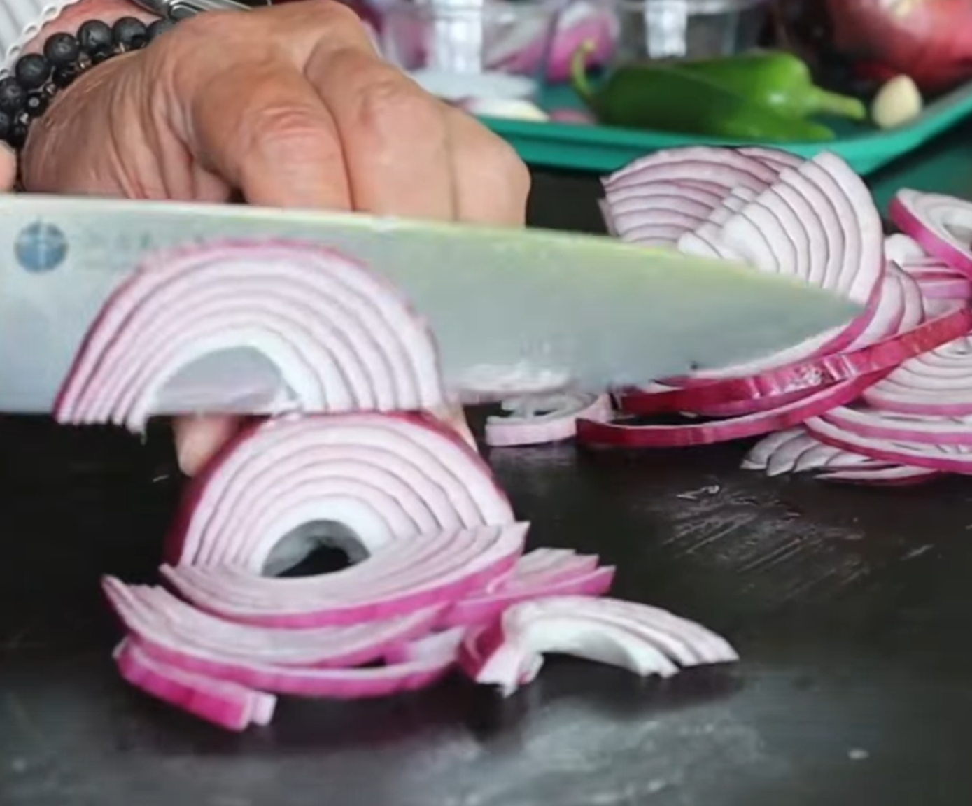 A hand slices a red onion with a knife on a dark surface. Thin onion rings are neatly cut, and more onions and vegetables are visible in the blurred background.