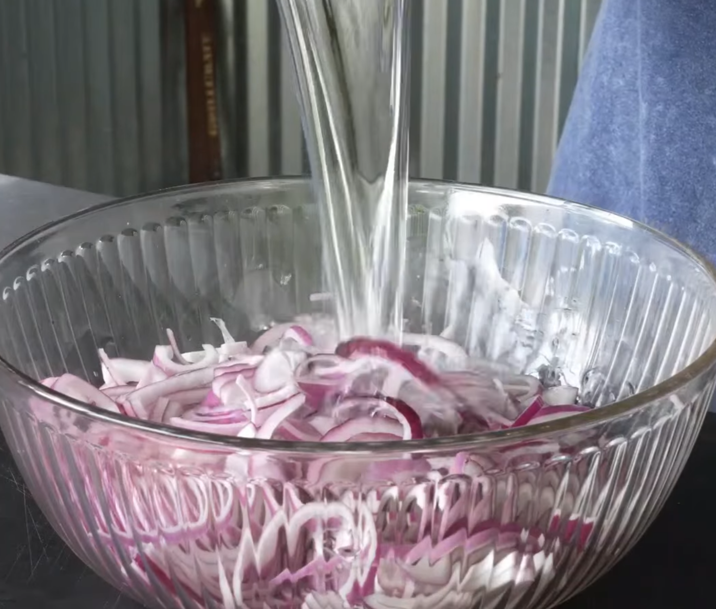 A clear bowl of sliced red onions is shown as water is being poured over them, with a metal surface and a blue cloth visible in the background.