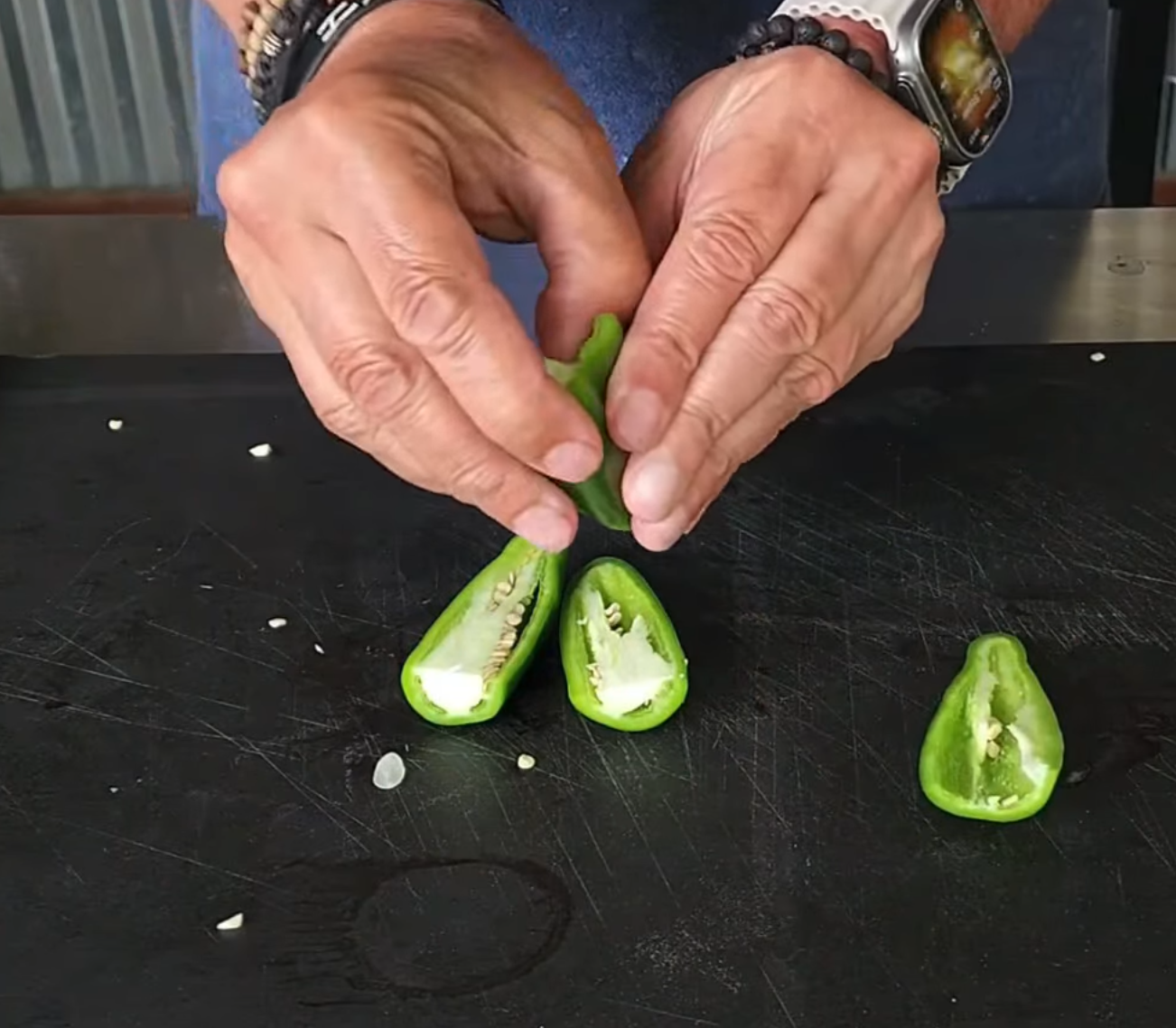 Two hands wearing bracelets and a watch are opening a halved jalapeño pepper on a black cutting board, with another halved pepper and scattered seeds nearby.