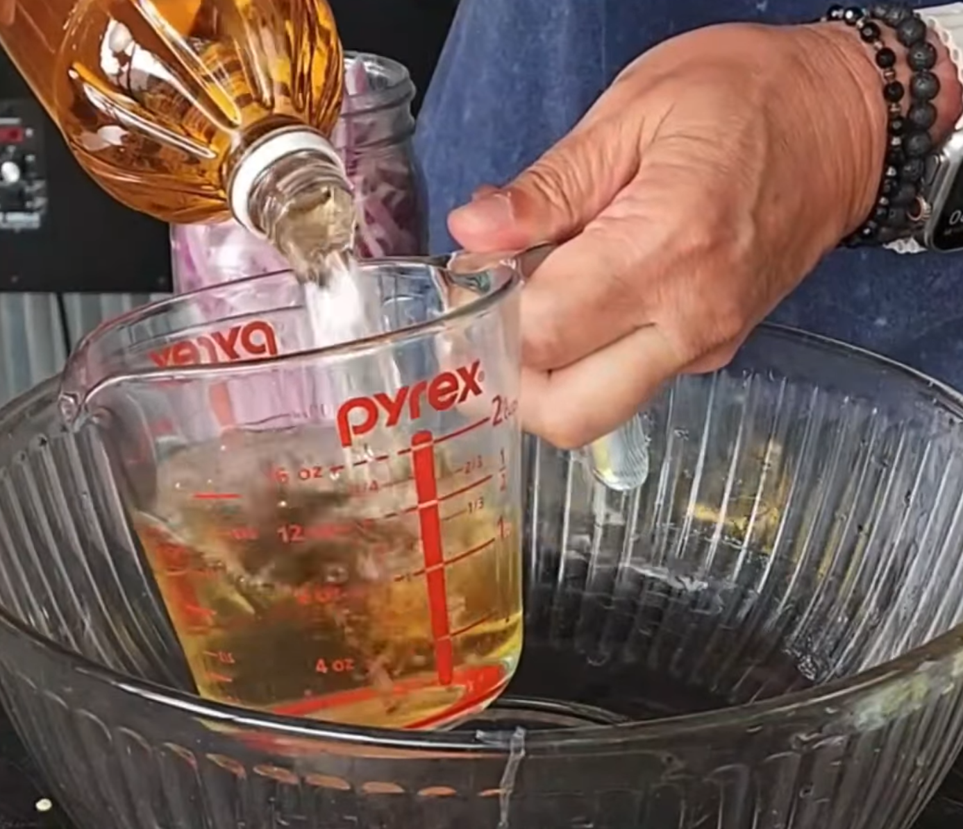 A person pours liquid from a bottle into a glass measuring cup. The cup is held over a large glass mixing bowl. The persons hands and part of their blue sleeve are visible.