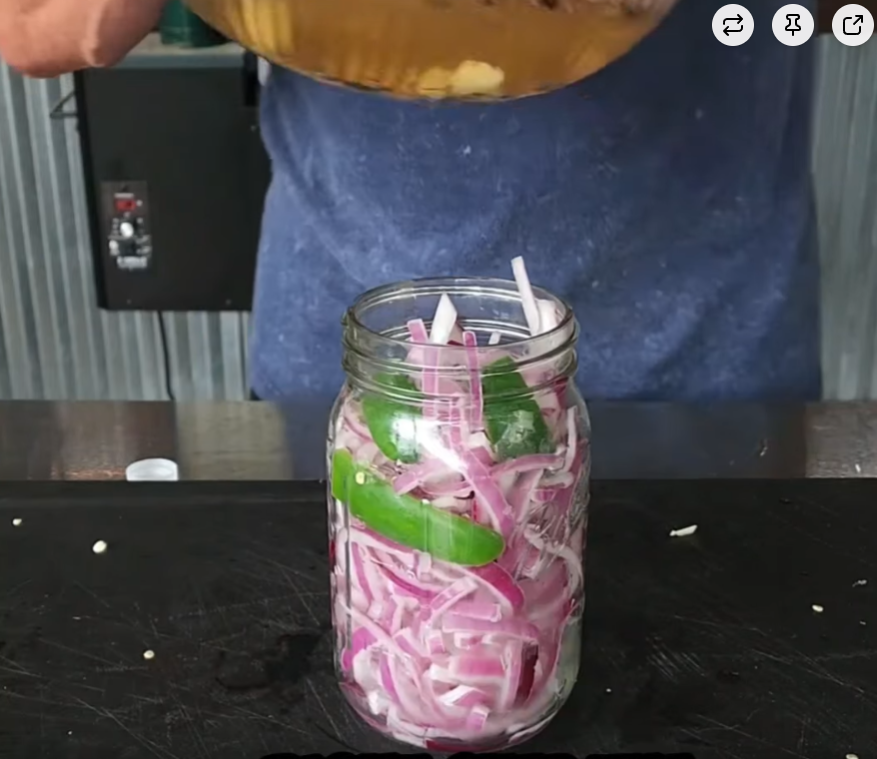 A person pours liquid into a glass jar filled with sliced red onions and green peppers. The jar sits on a black surface, and only the persons torso and arms are visible.