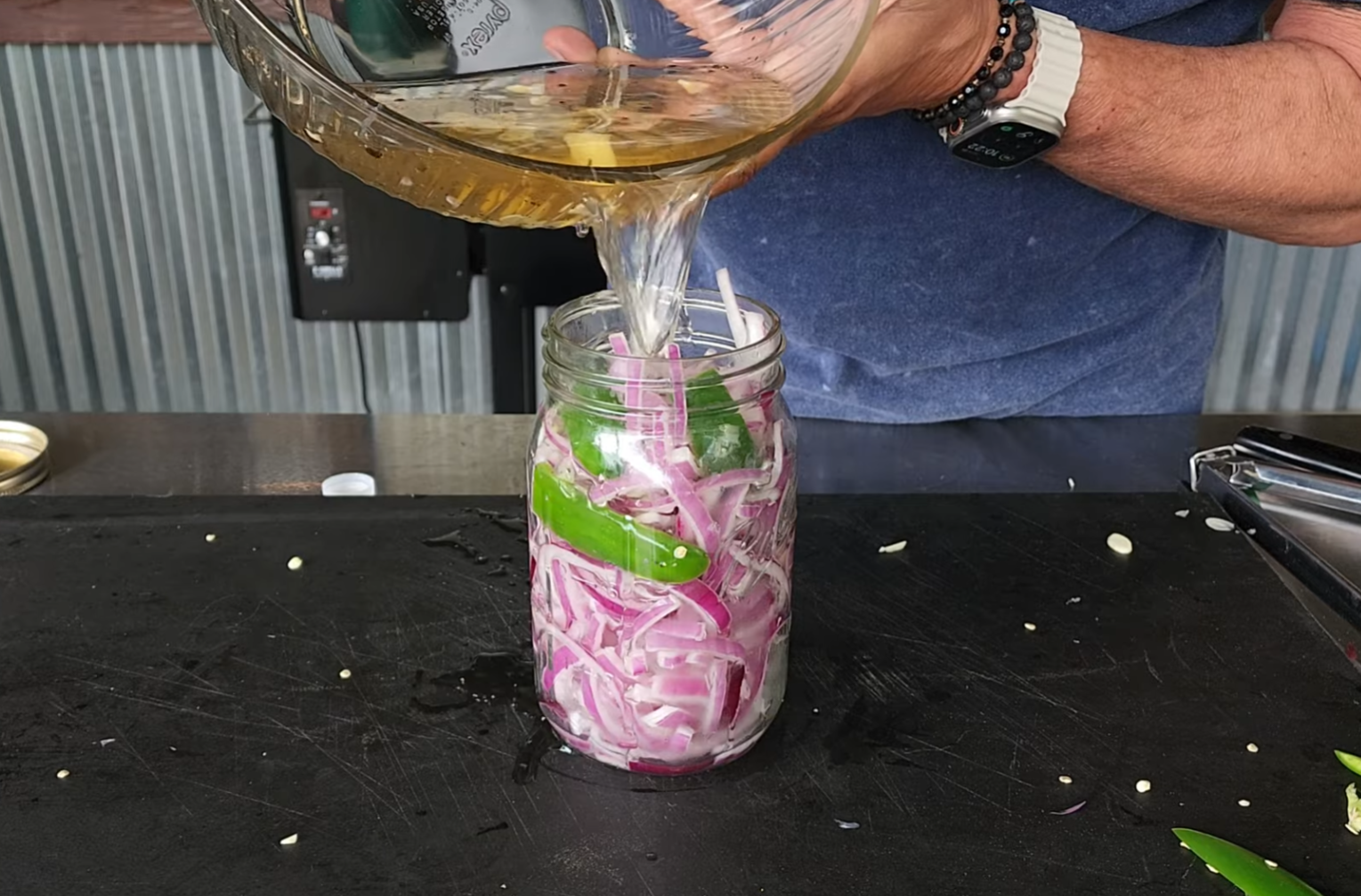 A person pours brine into a jar filled with sliced red onions and green peppers on a black countertop.