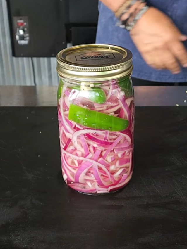 A jar filled with pickled red onions and a green chili pepper sits on a black surface, with a person in the background wearing a blue shirt.