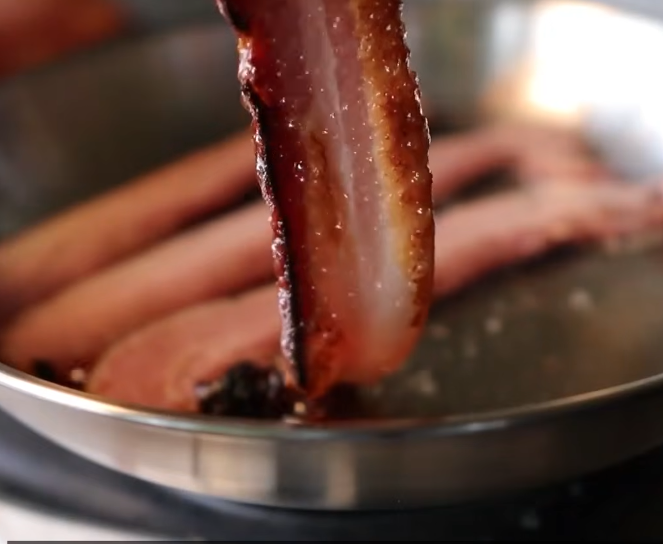 A crispy strip of bacon is held above a frying pan with other bacon pieces sizzling in hot grease.