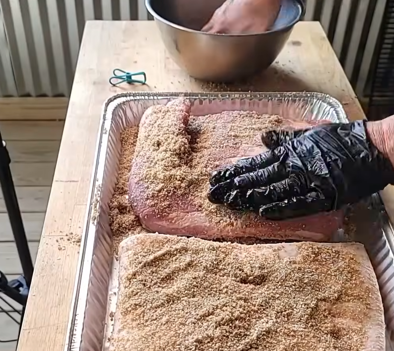 A person wearing black gloves rubs seasoning onto large cuts of meat in a foil tray. Another person mixes something in a metal bowl nearby on a wooden table.