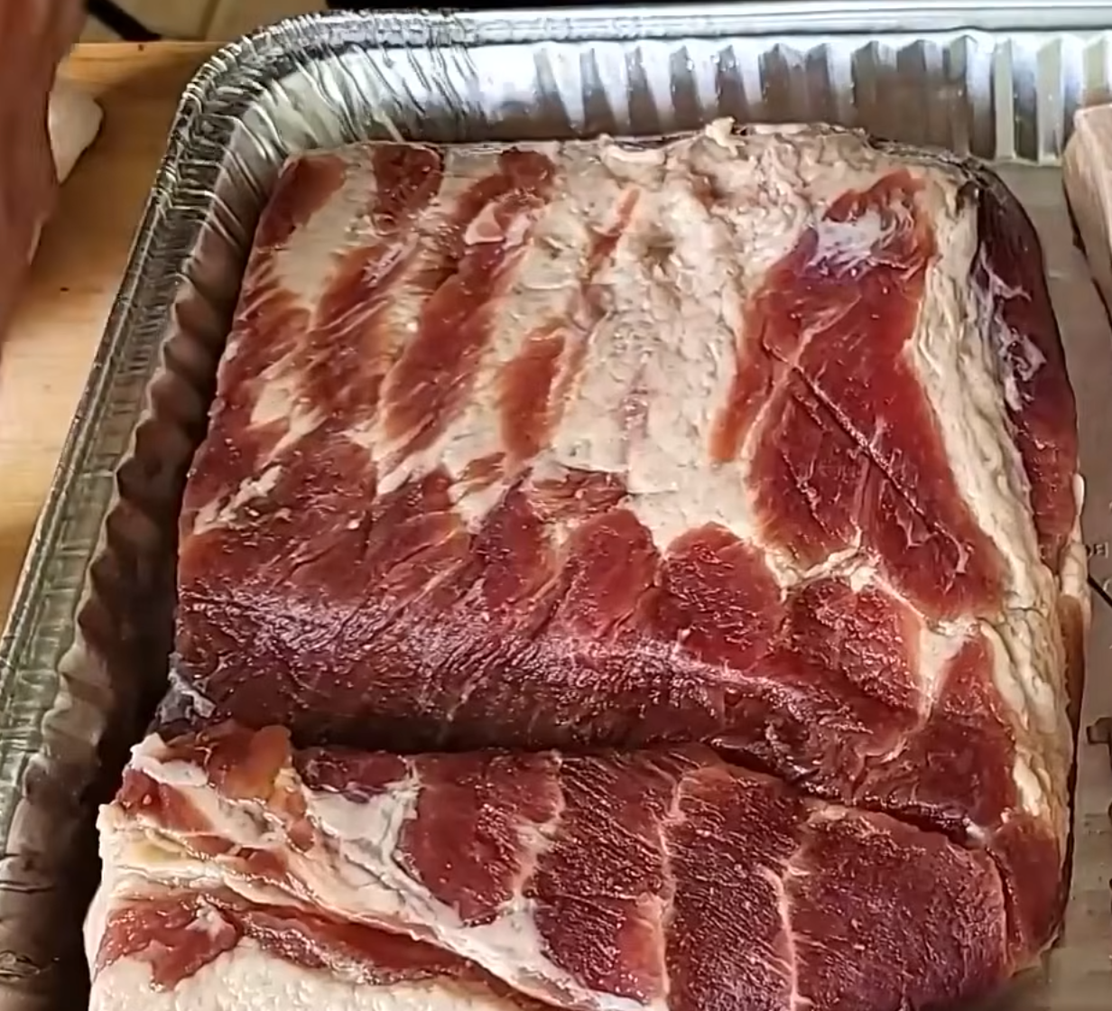 A large slab of raw pork belly with visible fat and meat layers sits in a metal tray on a wooden surface, ready for preparation.