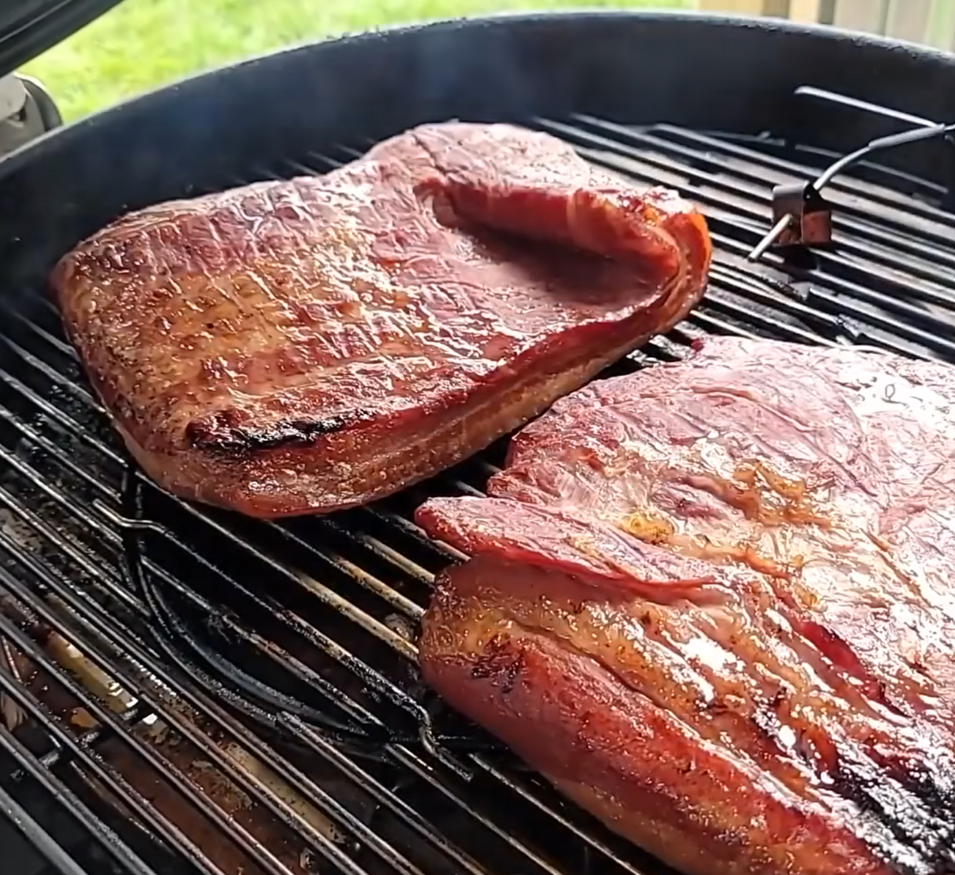 Two large slabs of brisket are smoking on a round grill outdoors, with visible grill marks and a reddish-brown, slightly glistening surface. Smoke rises from the grill, and grass is seen in the background.