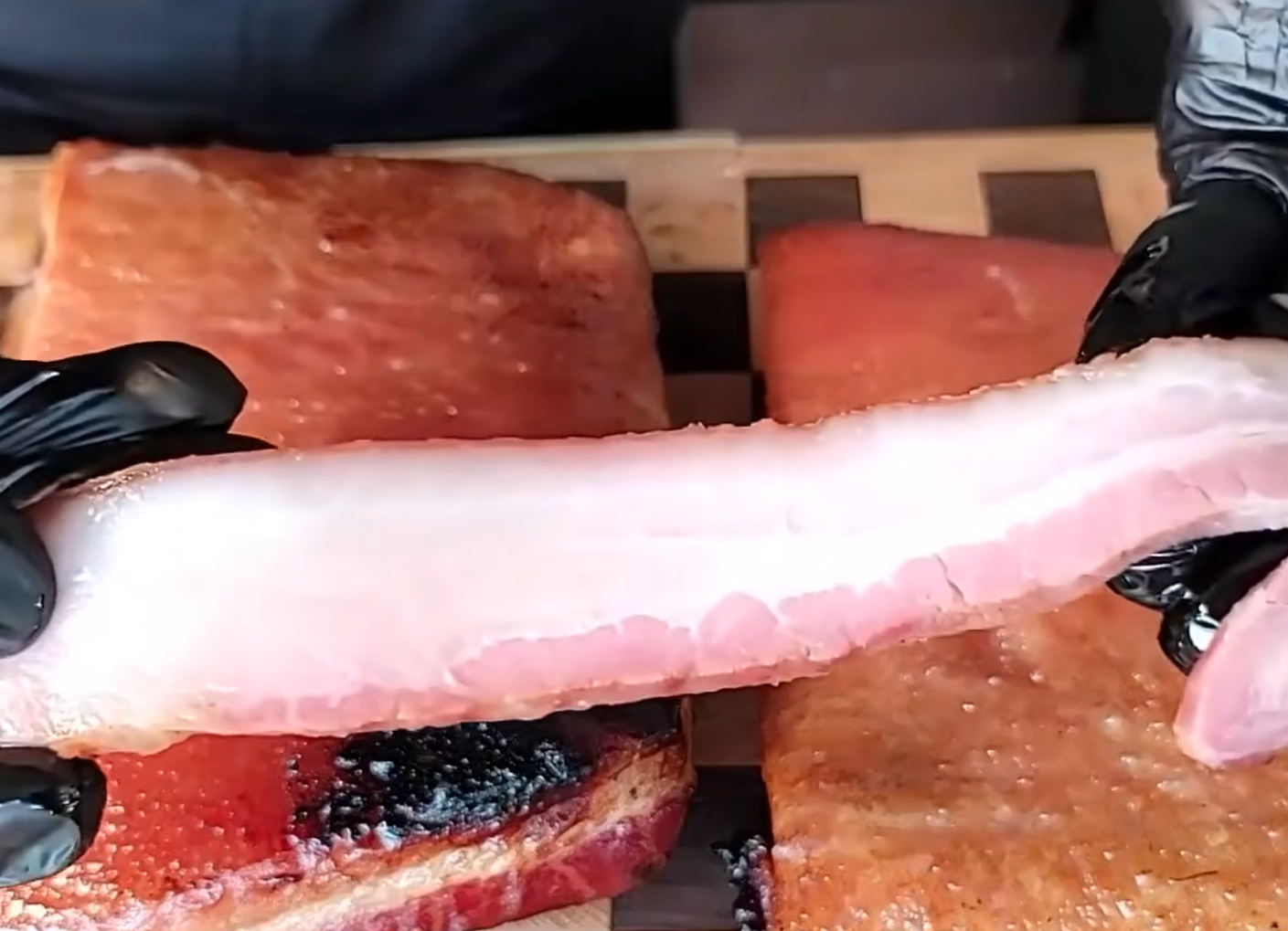 A person wearing black gloves holds a raw fish fillet over several cooked or smoked fish fillets that are resting on a wooden cutting board.