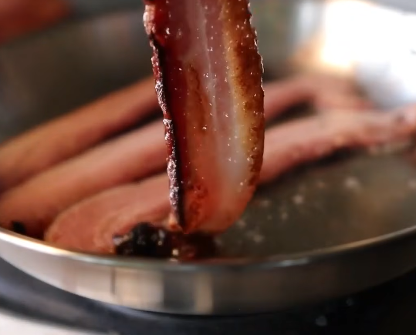 Close-up of crispy bacon being lifted from a frying pan with more strips cooking inside, showing sizzling grease and browned edges.
