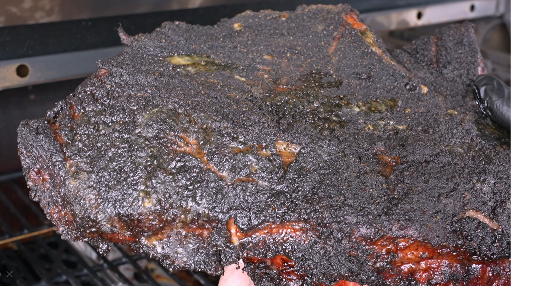 A large slab of smoked brisket with a dark, textured bark is being held over a grill, showing crispy edges and hints of rendered fat.