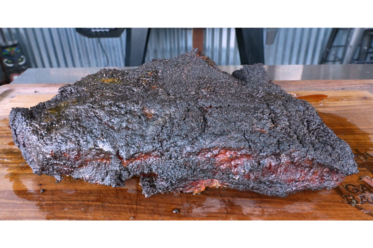 A large smoked brisket with a dark, crispy bark rests on a wooden cutting board, showing juicy meat and rendered fat.