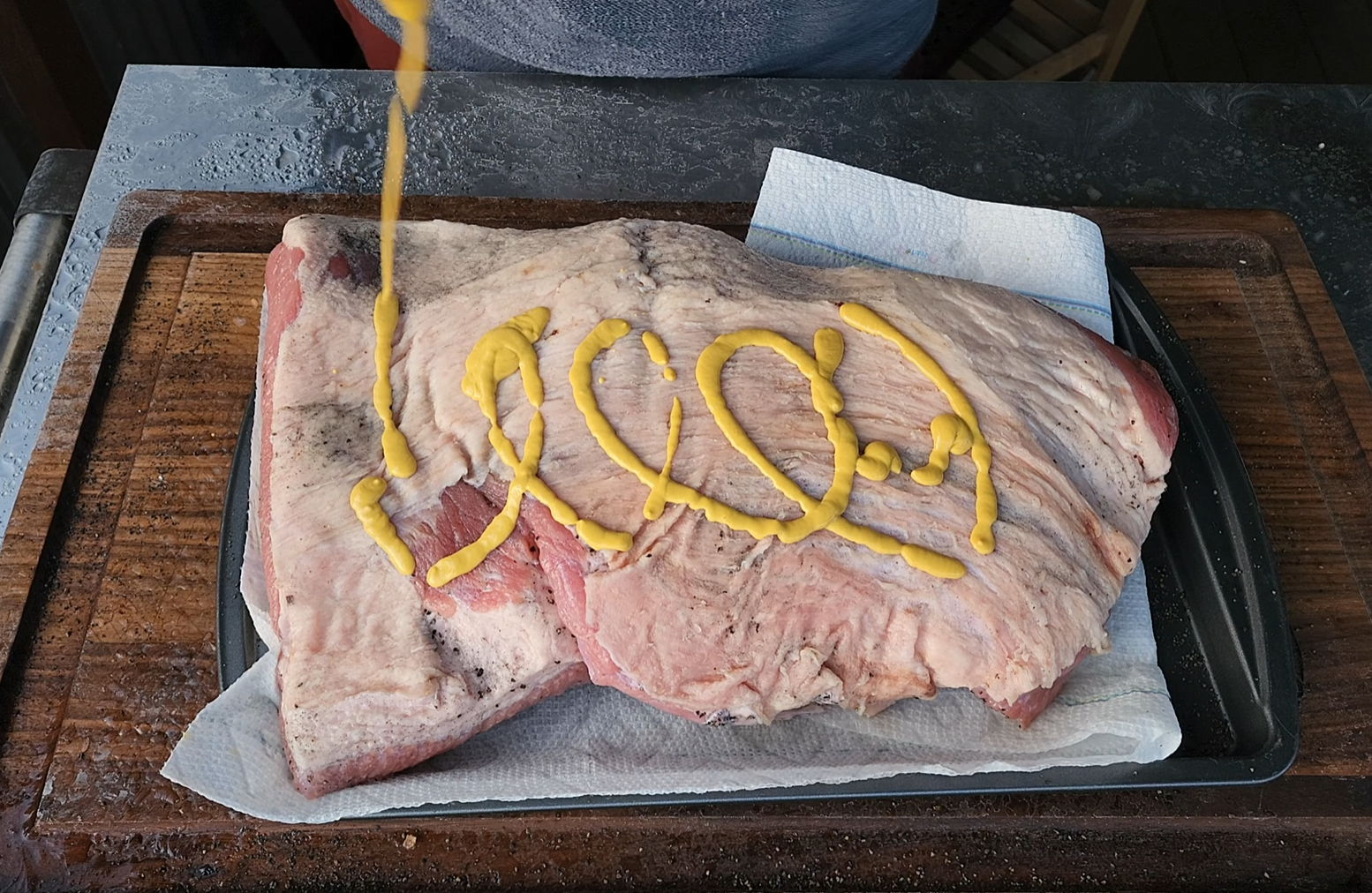 A slab of raw brisket on a tray lined with paper towels is being topped with a zigzag of yellow mustard.