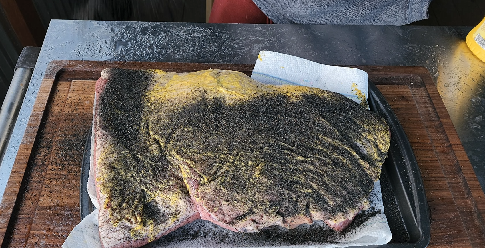 A large cut of raw meat on a wooden board is coated with yellow mustard and black pepper seasoning, resting on paper towels. A persons midsection is partially visible above the meat.