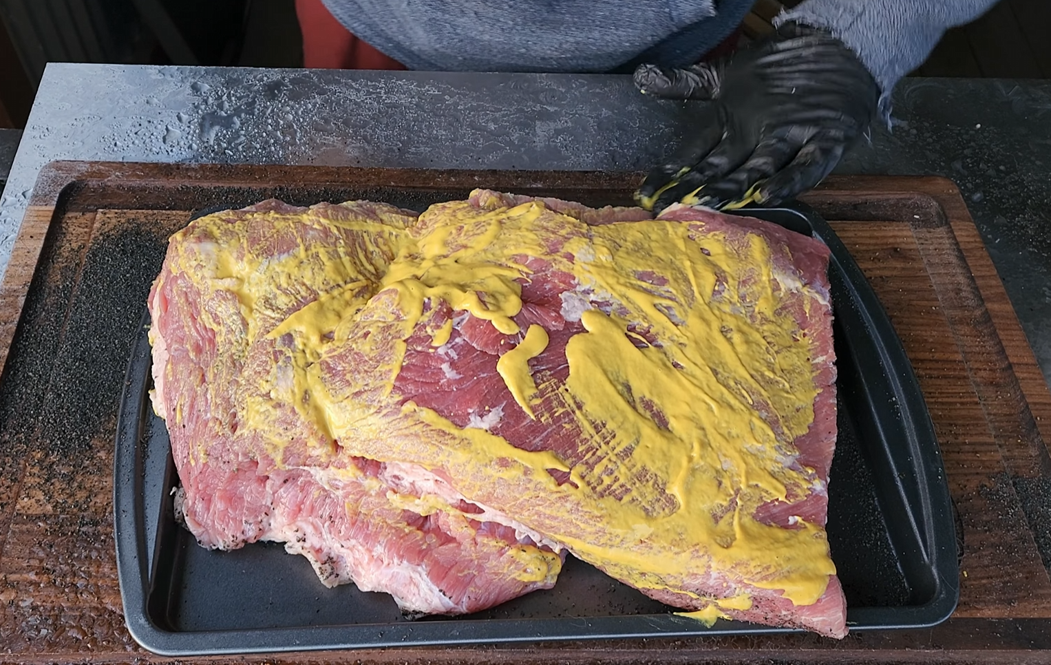 Two large pieces of raw brisket covered in yellow mustard are placed on a black tray, resting on a wooden cutting board. A person wearing a gray shirt and black glove is working beside the meat.
