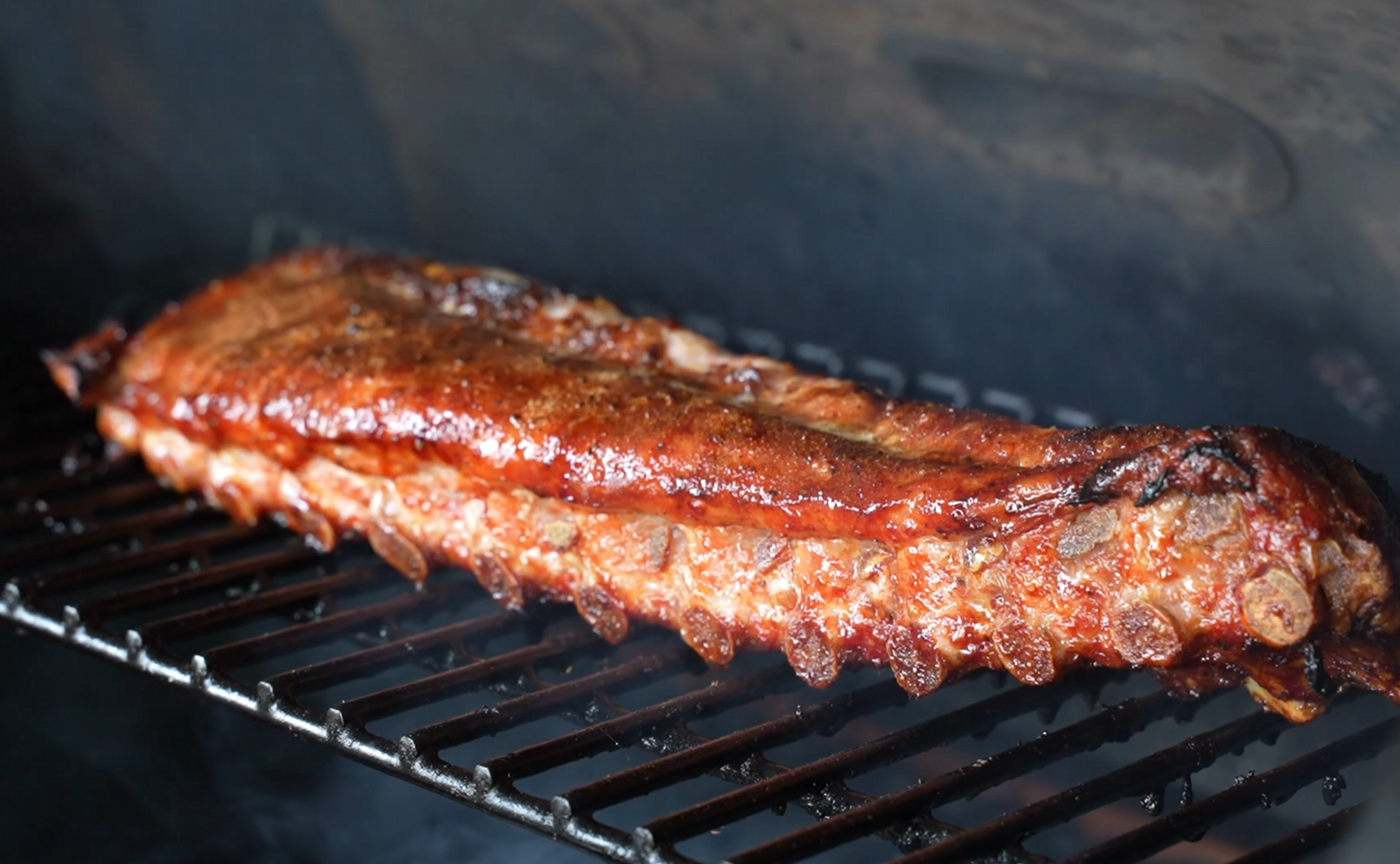 A rack of ribs with a glazed, caramelized surface cooks on a grill with visible smoke, highlighting the rich textures and savory color of the barbecued meat.