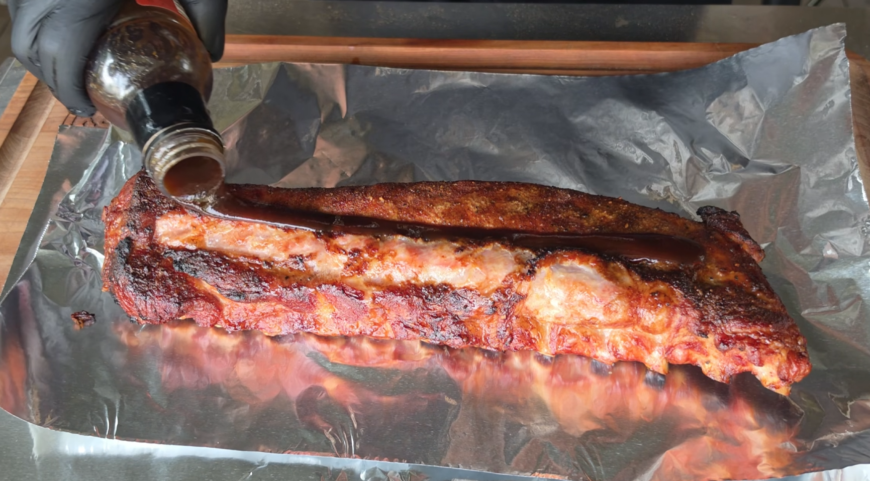 Barbecue sauce is being poured from a bottle onto a cooked rack of ribs resting on a sheet of aluminum foil.