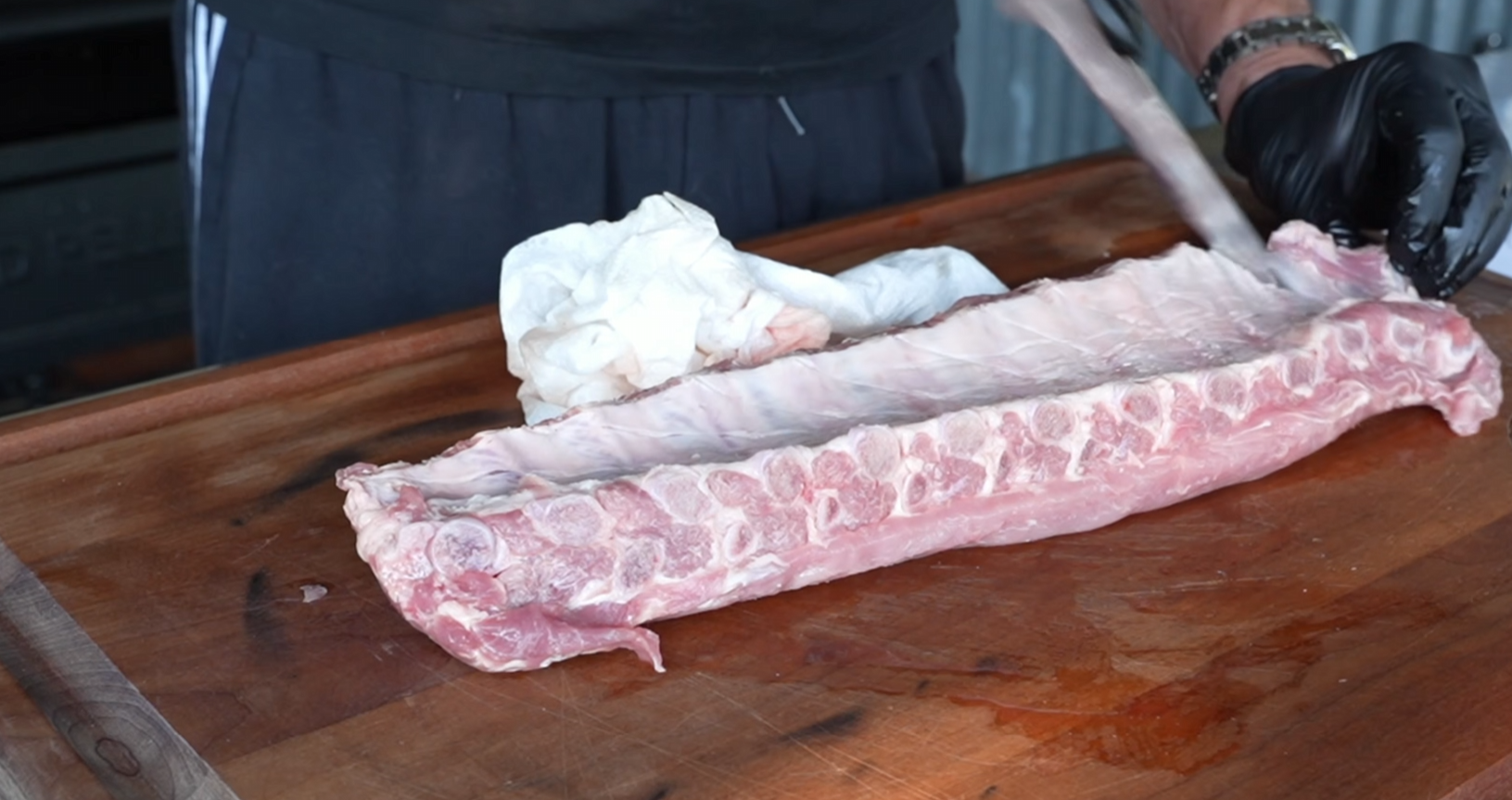 A person wearing a black glove uses a knife to remove the membrane from a raw rack of ribs on a wooden cutting board, with a paper towel nearby.