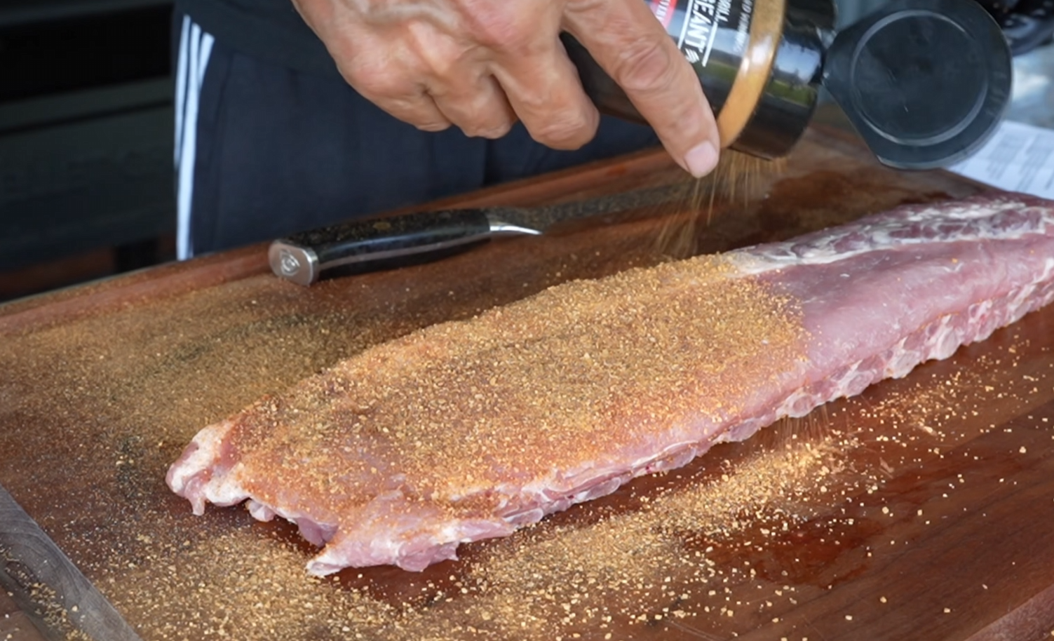 A person sprinkles seasoning onto a raw rack of pork ribs on a wooden cutting board, with a knife lying beside the meat.