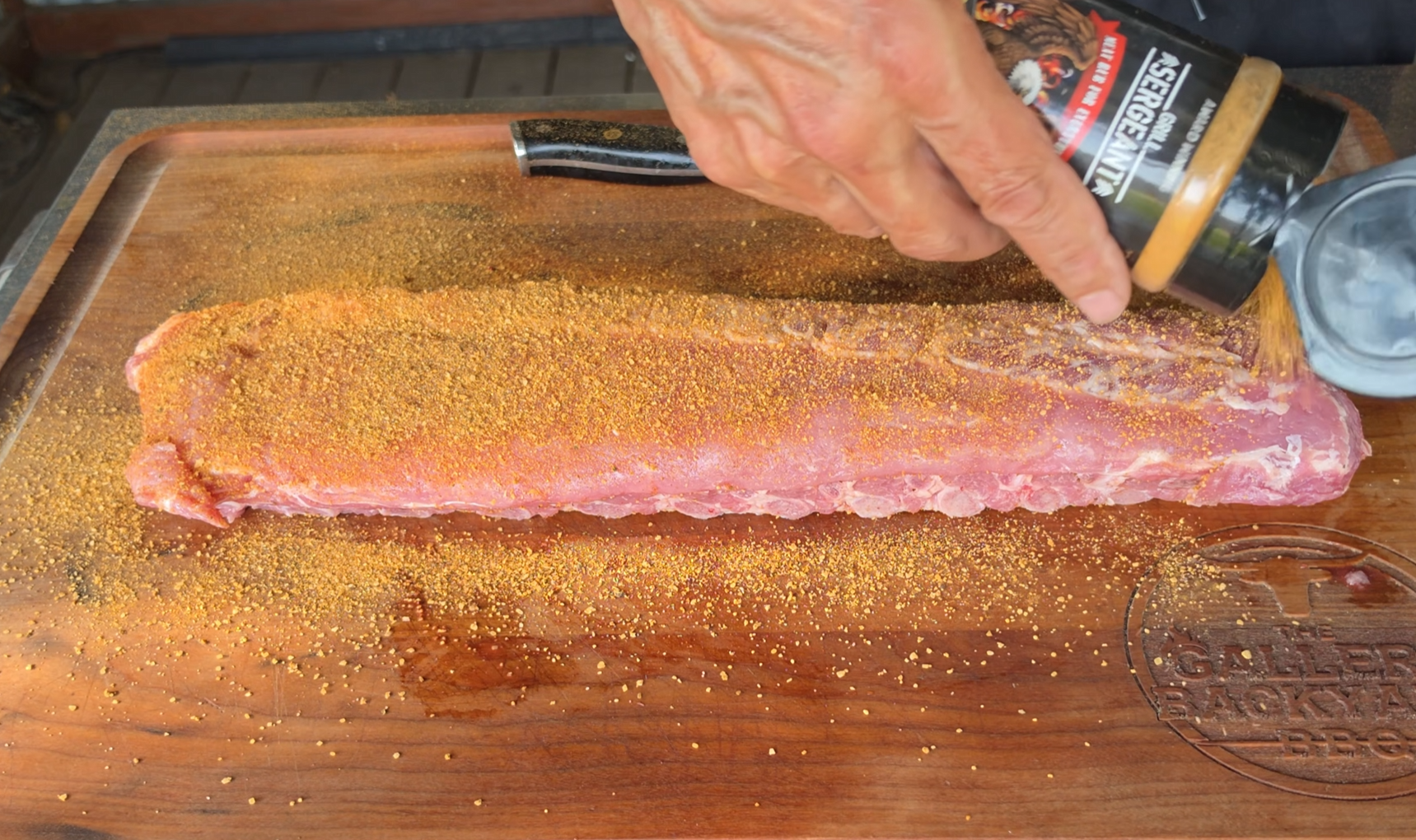 A hand sprinkles seasoning from a bottle onto a raw rack of pork ribs on a wooden cutting board engraved with a logo.
