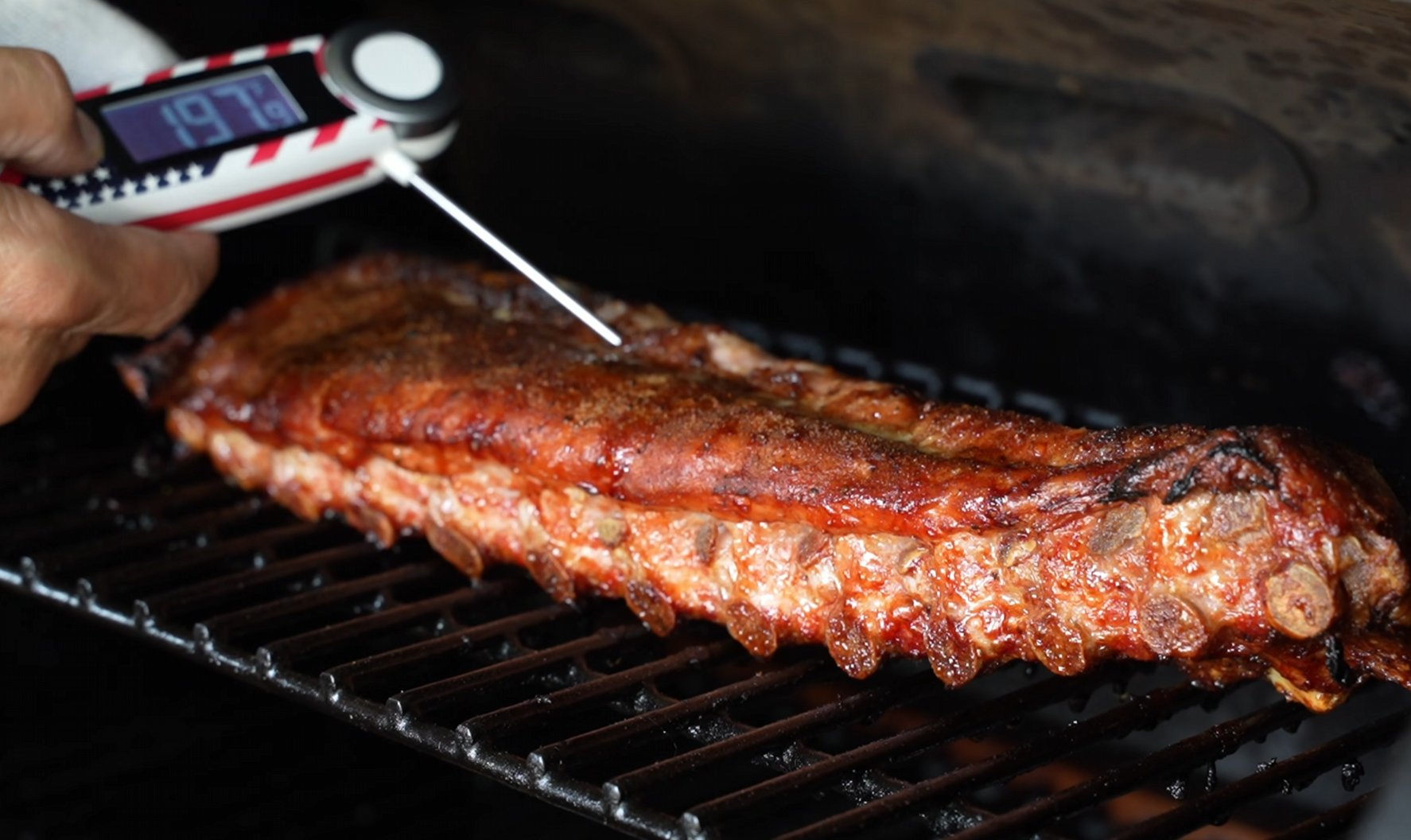 A rack of cooked ribs on a grill is checked for doneness with a meat thermometer, which reads 197.3°F.