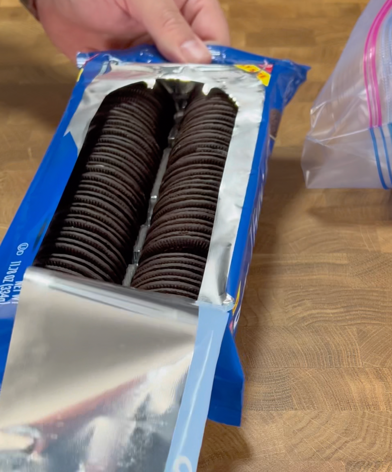 A hand holds an open package of chocolate sandwich cookies with white cream filling on a wooden surface. A resealable plastic bag sits nearby.