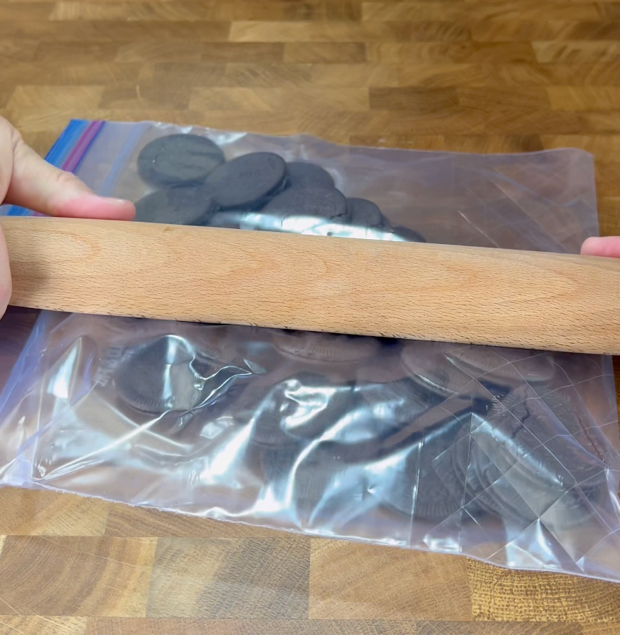 A person uses a wooden rolling pin to crush chocolate sandwich cookies inside a sealed plastic bag on a wooden surface.