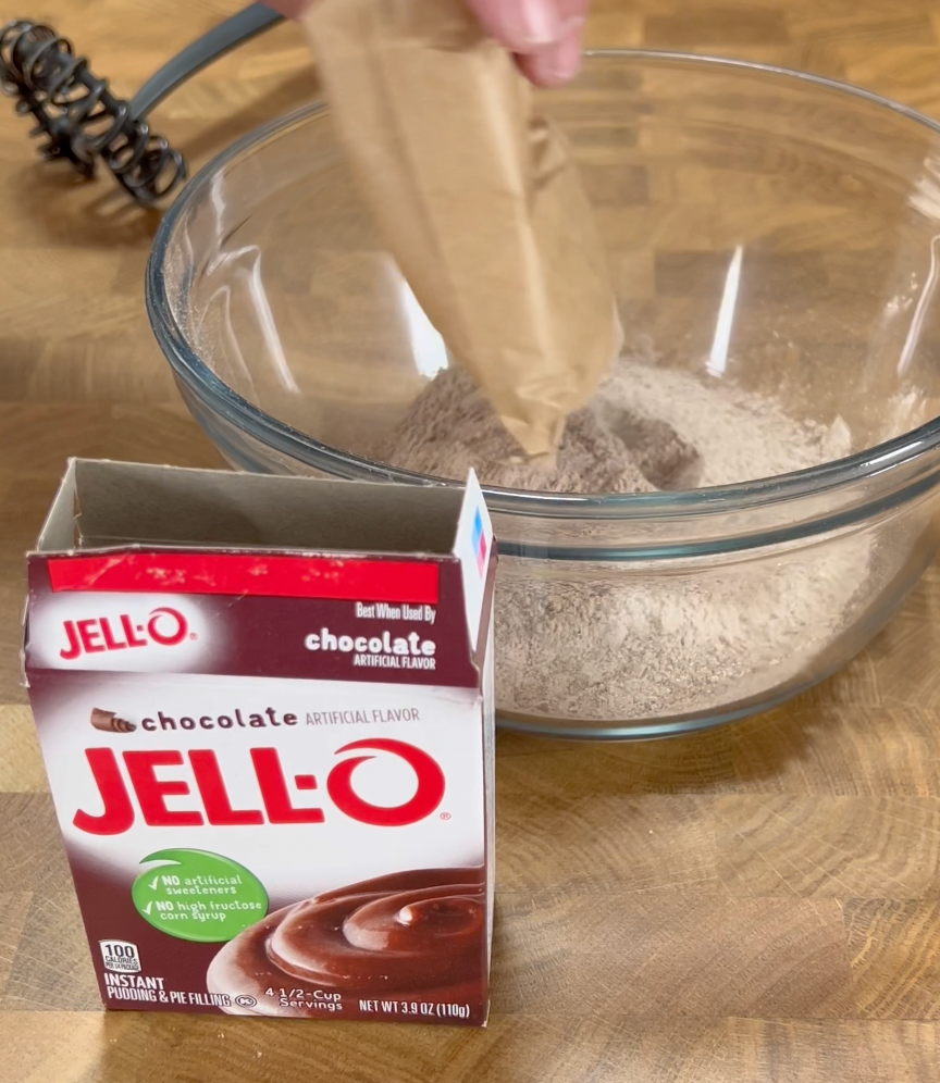A hand pours chocolate Jell-O pudding mix from a packet into a glass bowl. An empty Jell-O chocolate pudding mix box is on the table next to the bowl.