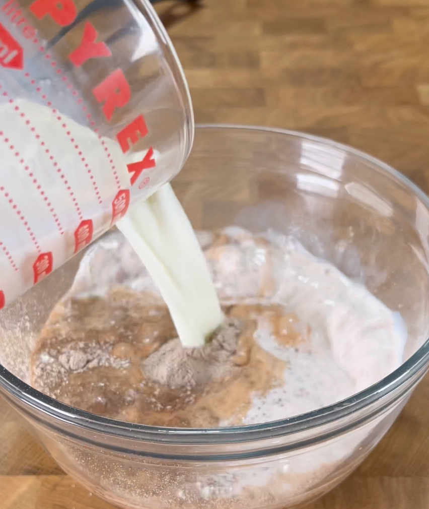 A glass measuring cup pours milk into a clear bowl containing baking ingredients like cocoa powder and flour on a wooden surface.