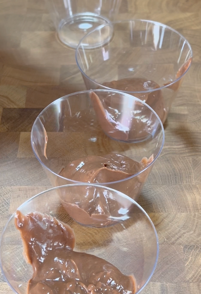 Three clear plastic cups with chocolate pudding dolloped inside are arranged on a wooden surface. An empty cup is visible in the background.