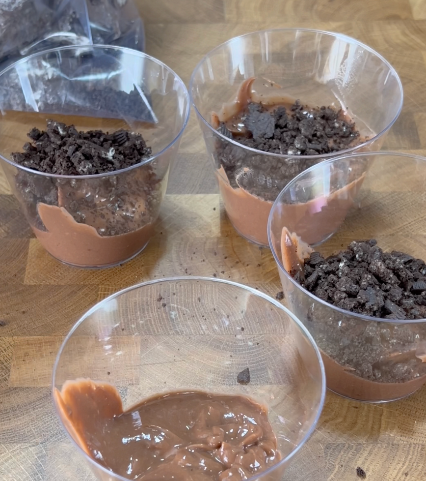 Four clear cups on a wooden surface, three filled with chocolate pudding topped with crushed cookies, and one with only chocolate pudding.