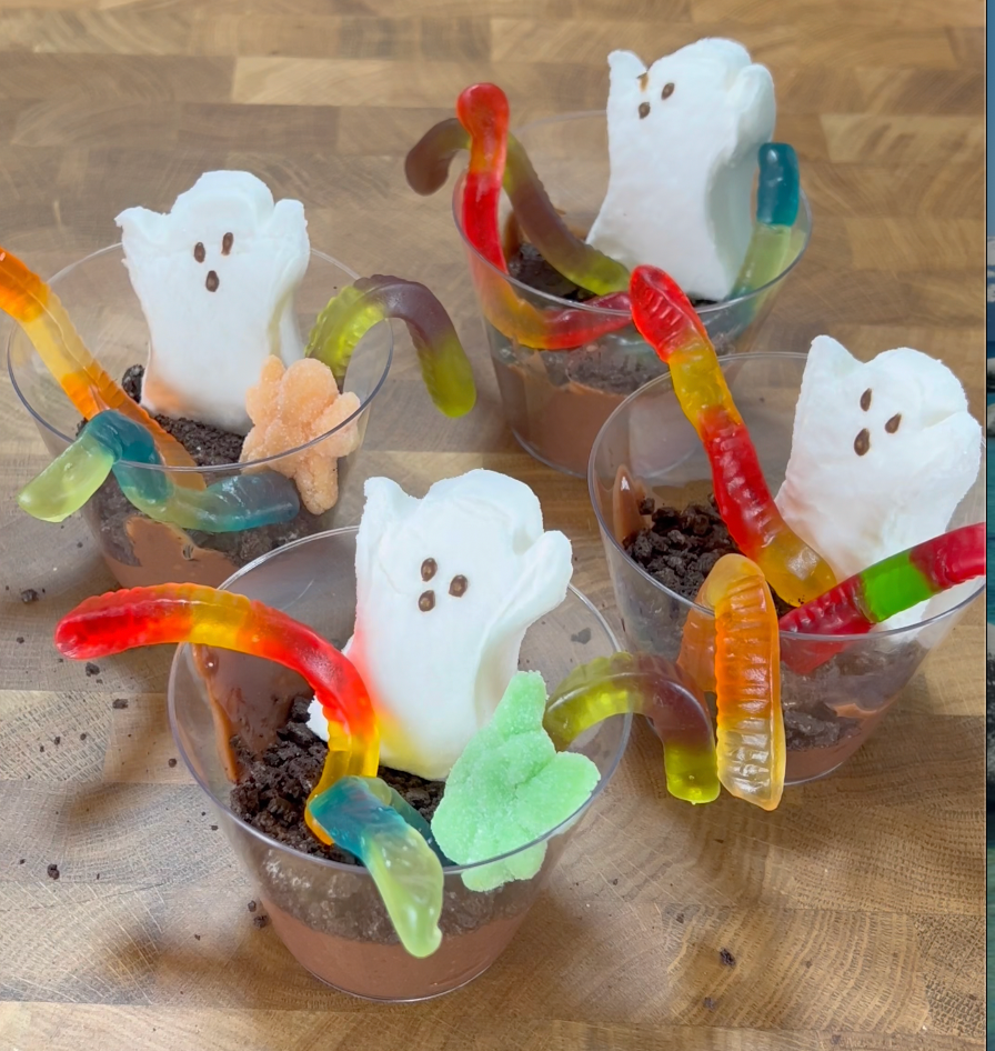 Four cups of chocolate pudding topped with crushed cookies, marshmallow ghosts, and gummy worms are arranged on a wooden surface, resembling a spooky Halloween-themed dessert.