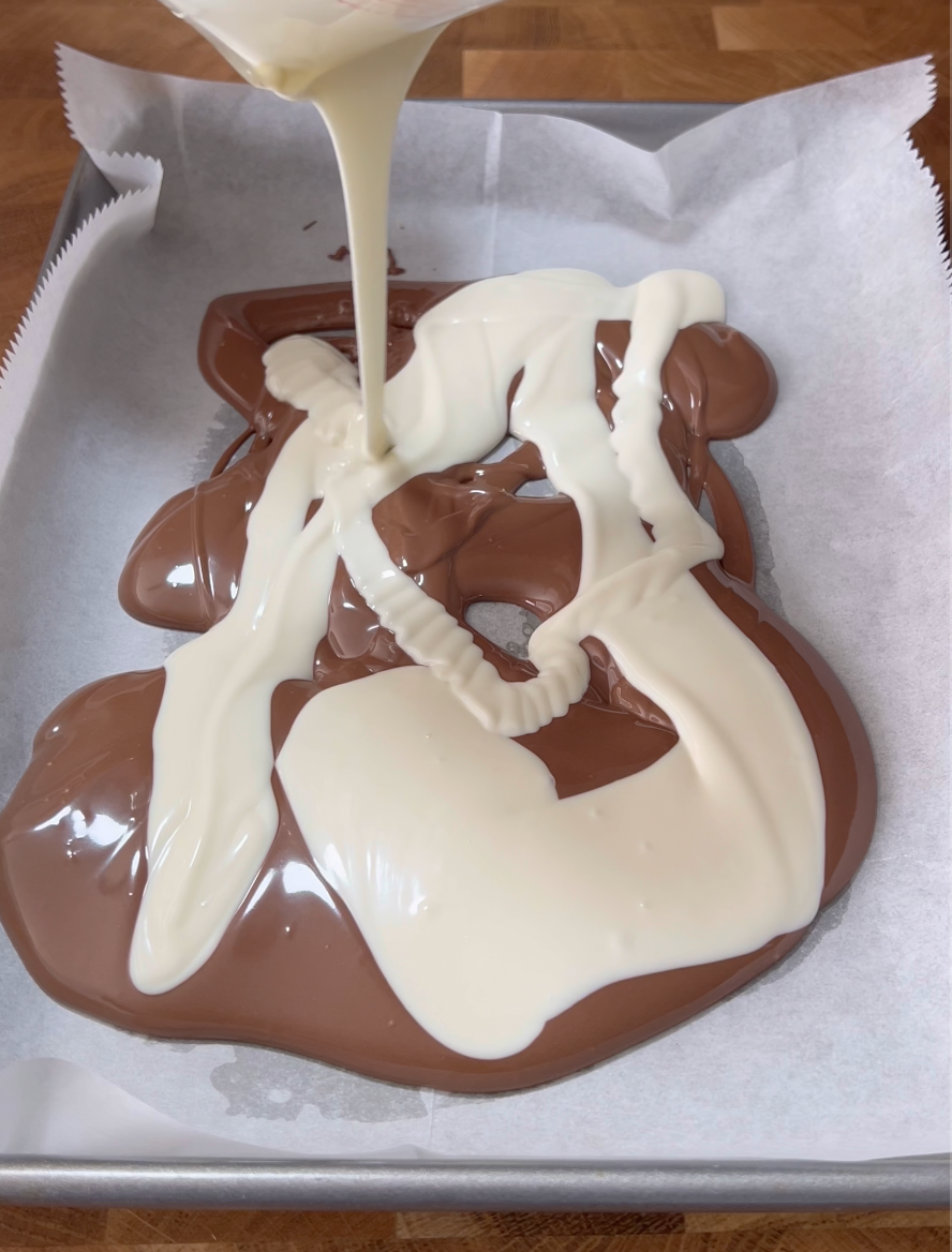Milk chocolate is spread on parchment paper in a baking tray, while melted white chocolate is being poured on top in swirls, creating a marbled effect, on a wooden surface.