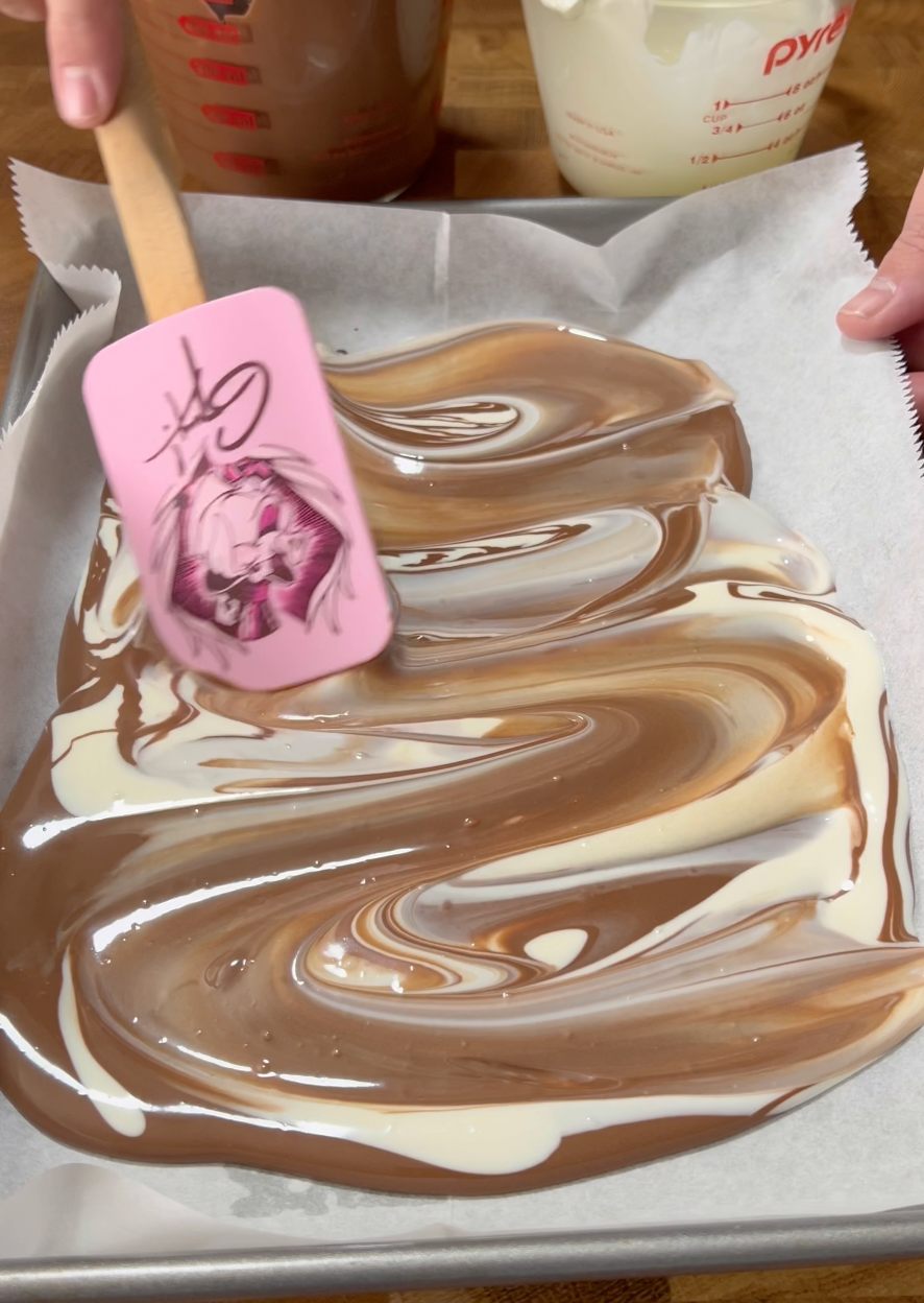 A hand spreads swirled milk, dark, and white chocolate on parchment paper in a pan using a pink spatula with an anime character illustration on it.