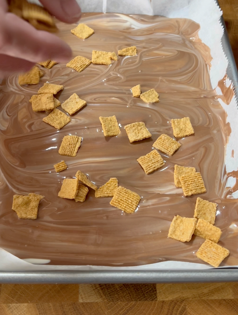 A hand sprinkles square cereal pieces onto a tray coated with melted chocolate, ready for a dessert preparation.