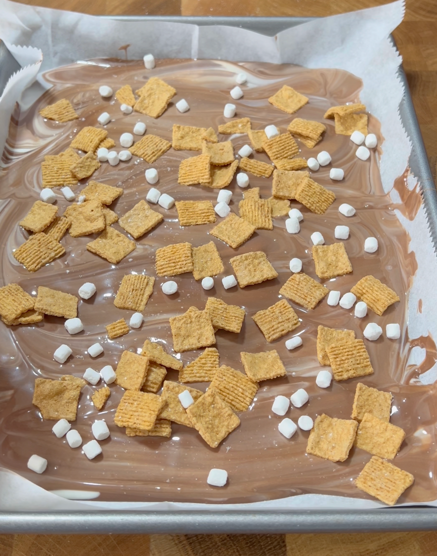 A baking pan lined with parchment paper holds a layer of melted chocolate, topped with golden cereal squares and mini marshmallows.